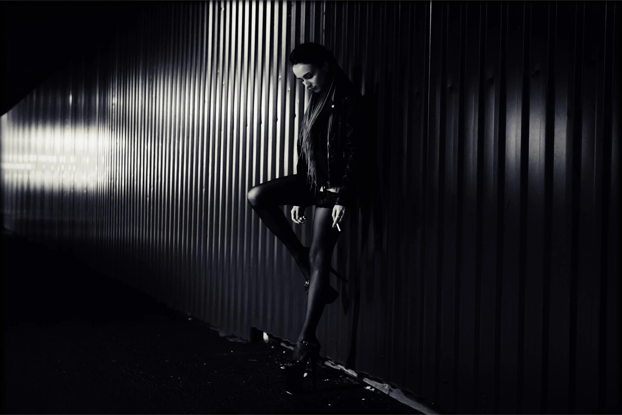 Woman in a leather jacket leaning against a metal wall in dim lighting, embodying a moody, edgy street style.
