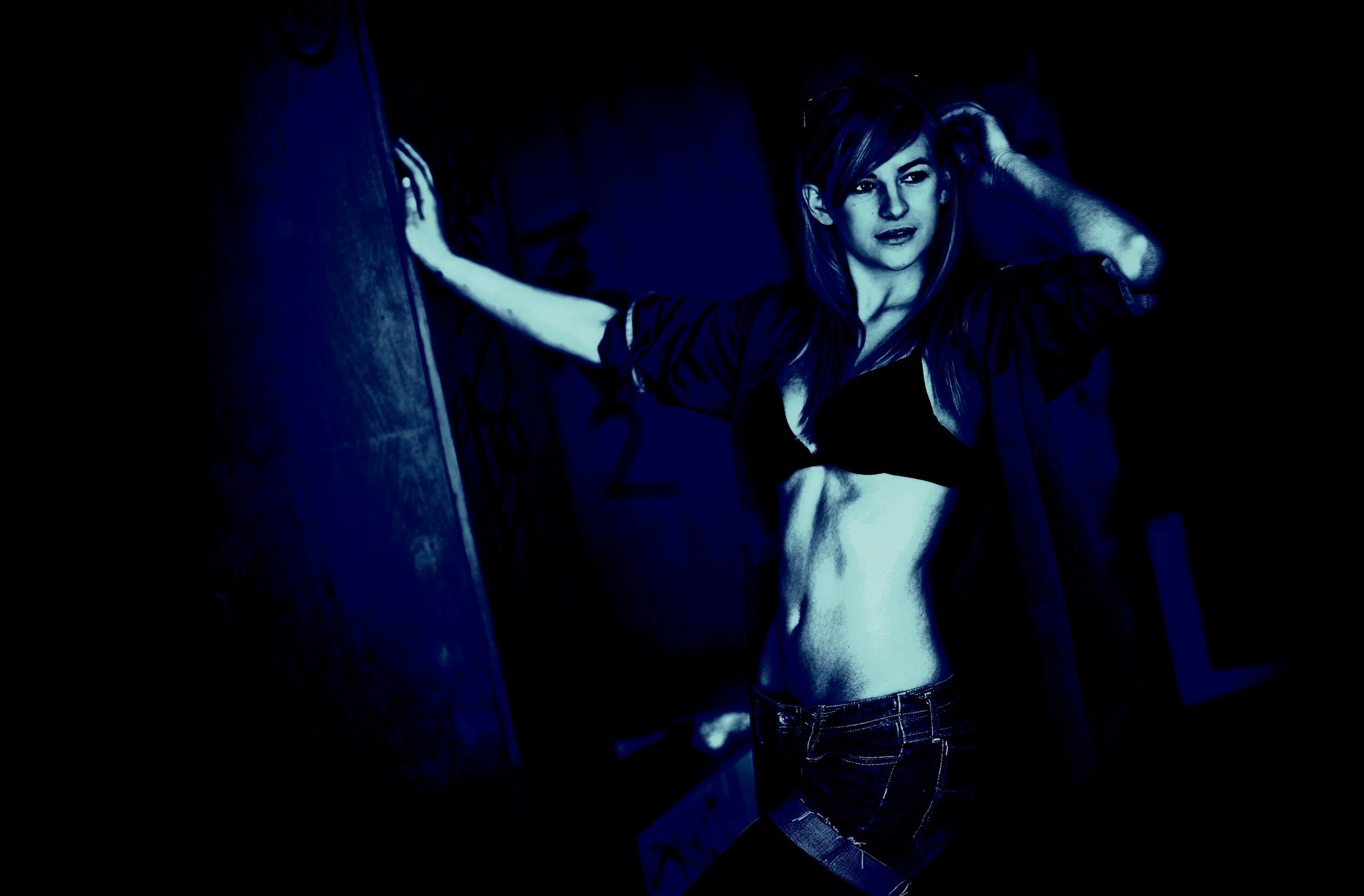 Woman in a dark urban setting, wearing a bra and open jacket, with jeans.