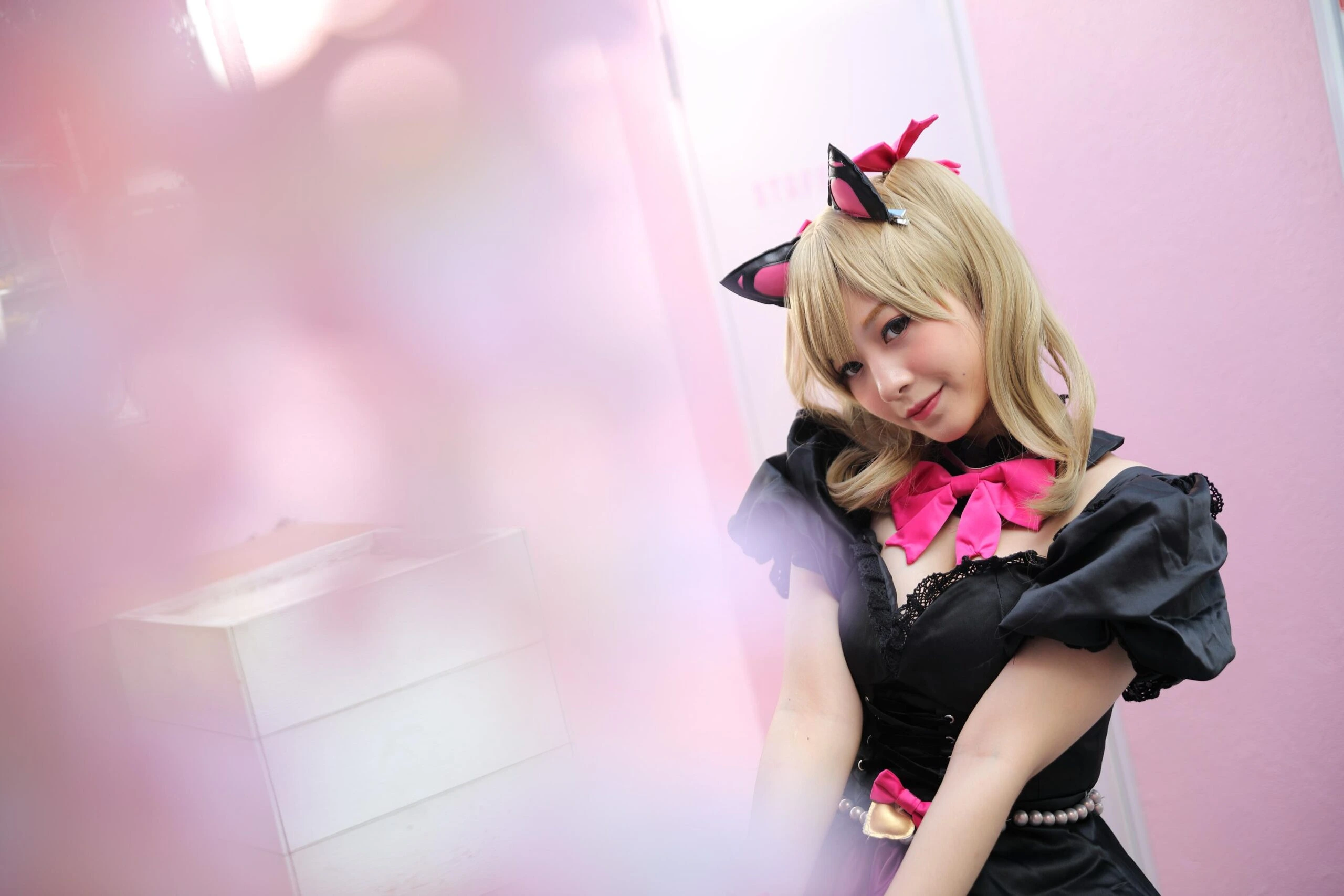 A cosplayer with blonde hair, dressed in a cute cat-themed outfit with black cat ears, a pink bow, and a black dress, posing in a soft, pastel-colored environment.