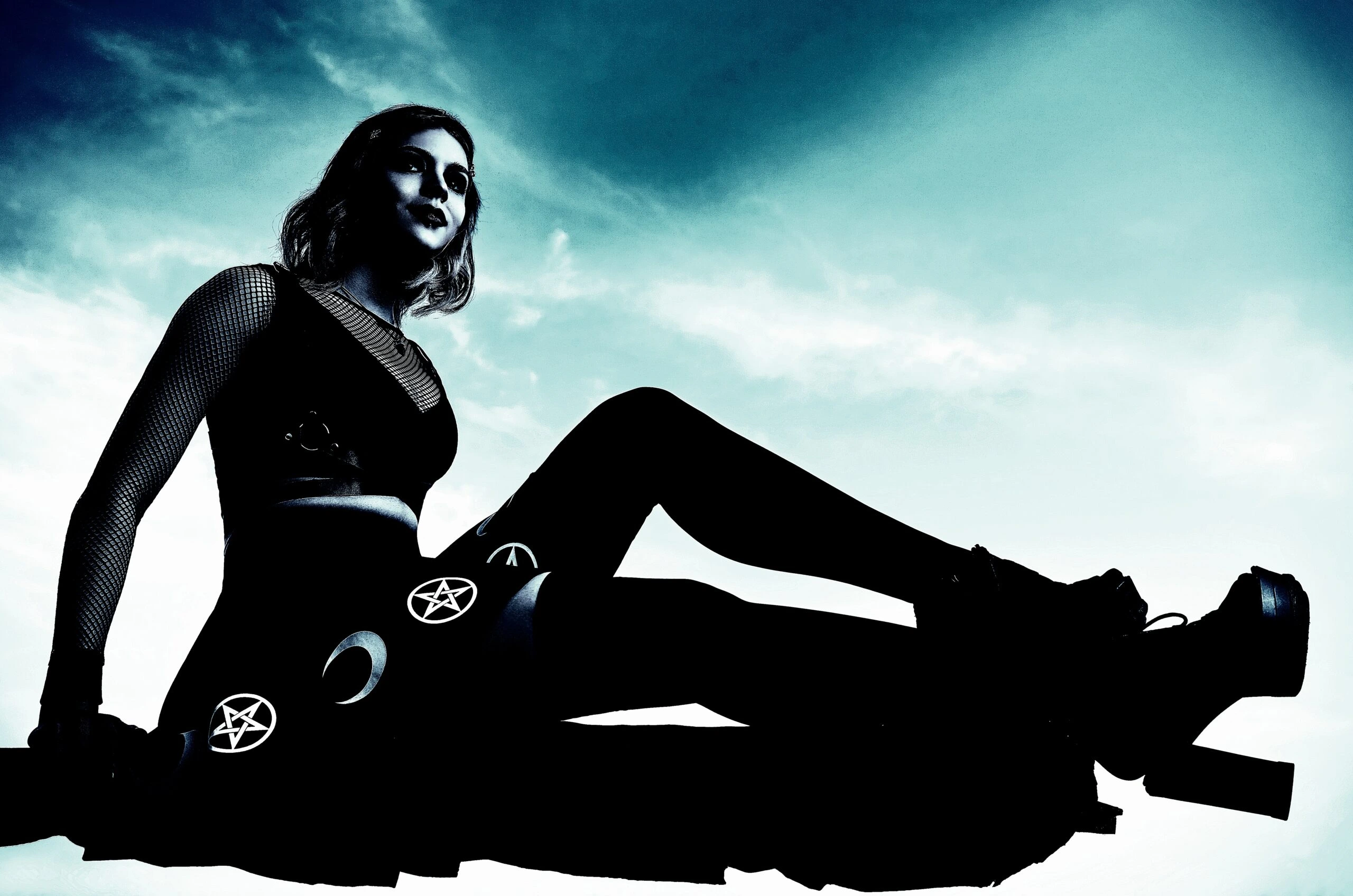 A woman in a gothic outfit sitting on a rooftop, wearing black stockings with pentagram symbols, under a cloudy sky.