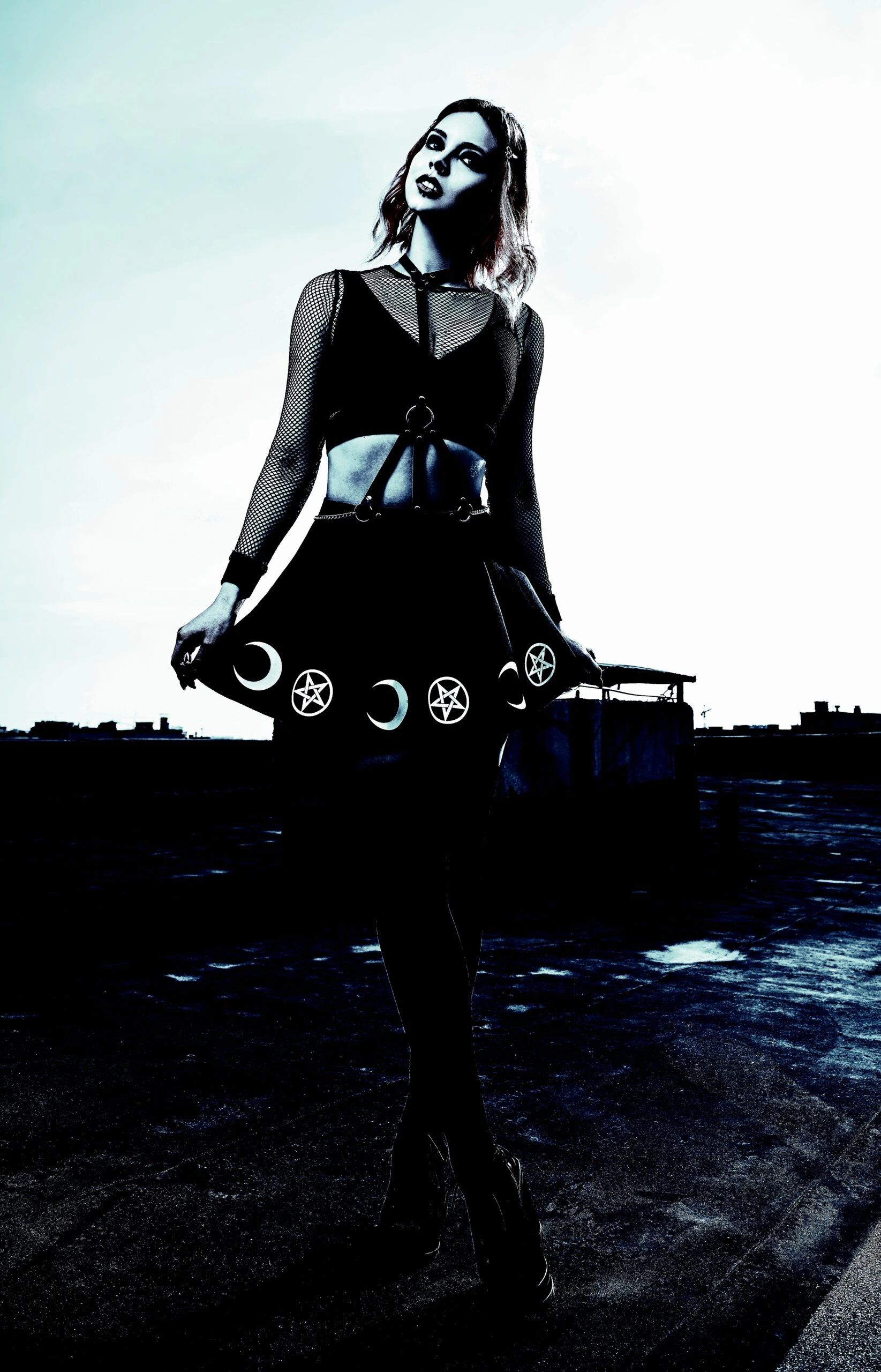 A woman in a gothic outfit standing on a rooftop, wearing a black skirt adorned with pentagram and moon symbols, against a cloudy sky.