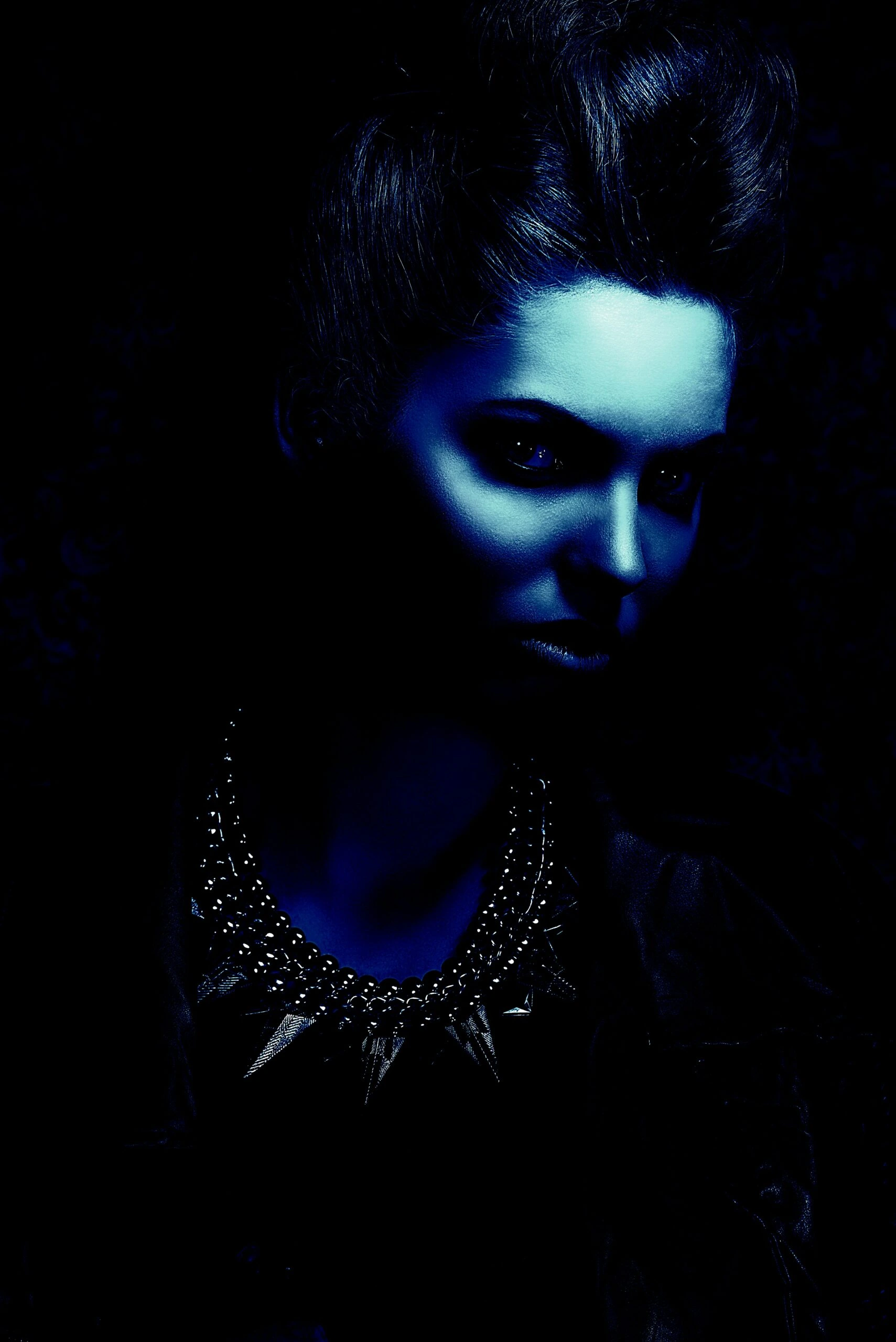 Woman with an intense gaze wearing a spiked gothic necklace under dim blue lighting, exuding dark elegance and mystery.