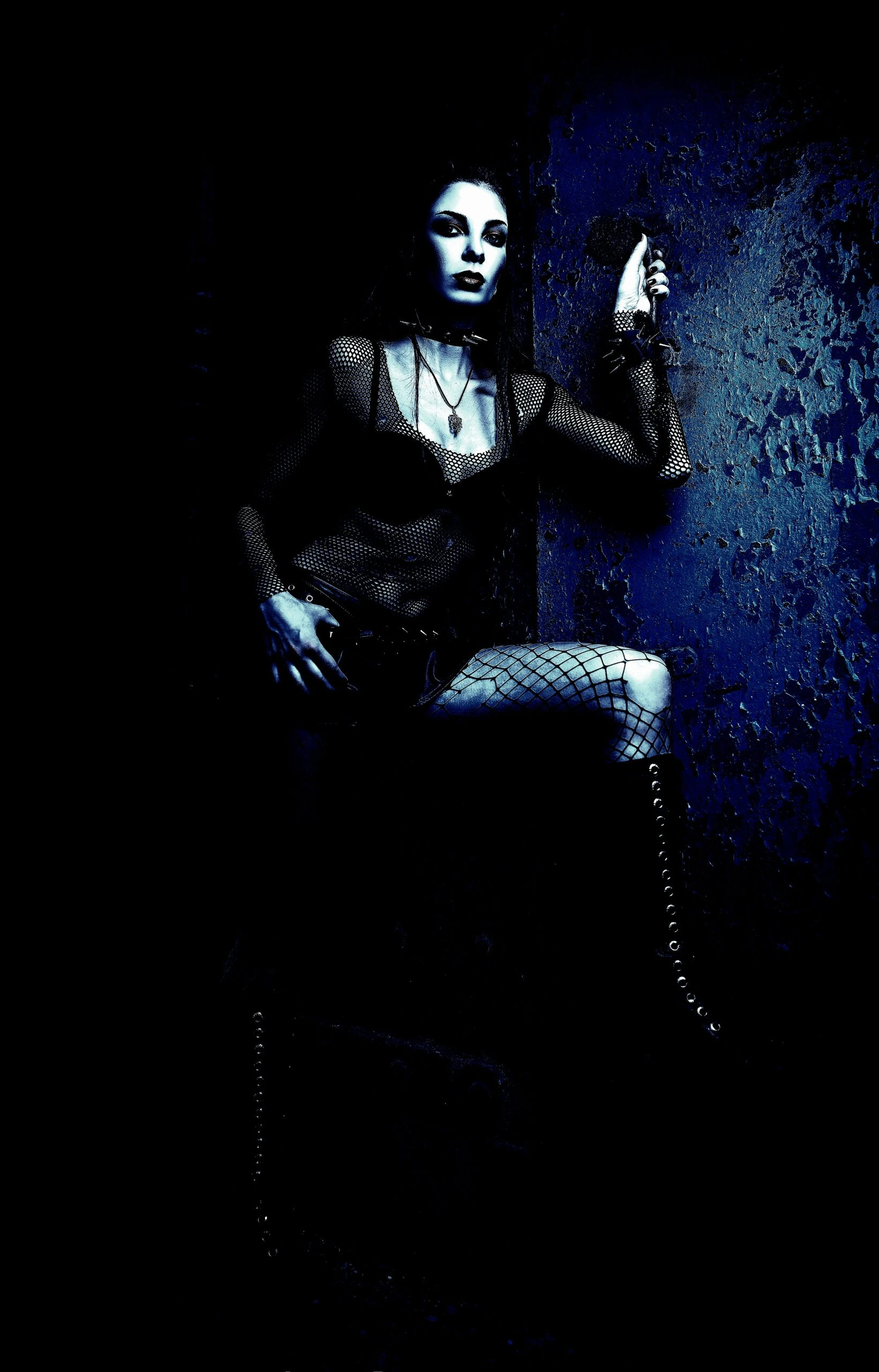 Gothic woman in a fishnet top and mesh sleeves, posing in a dark, textured environment.