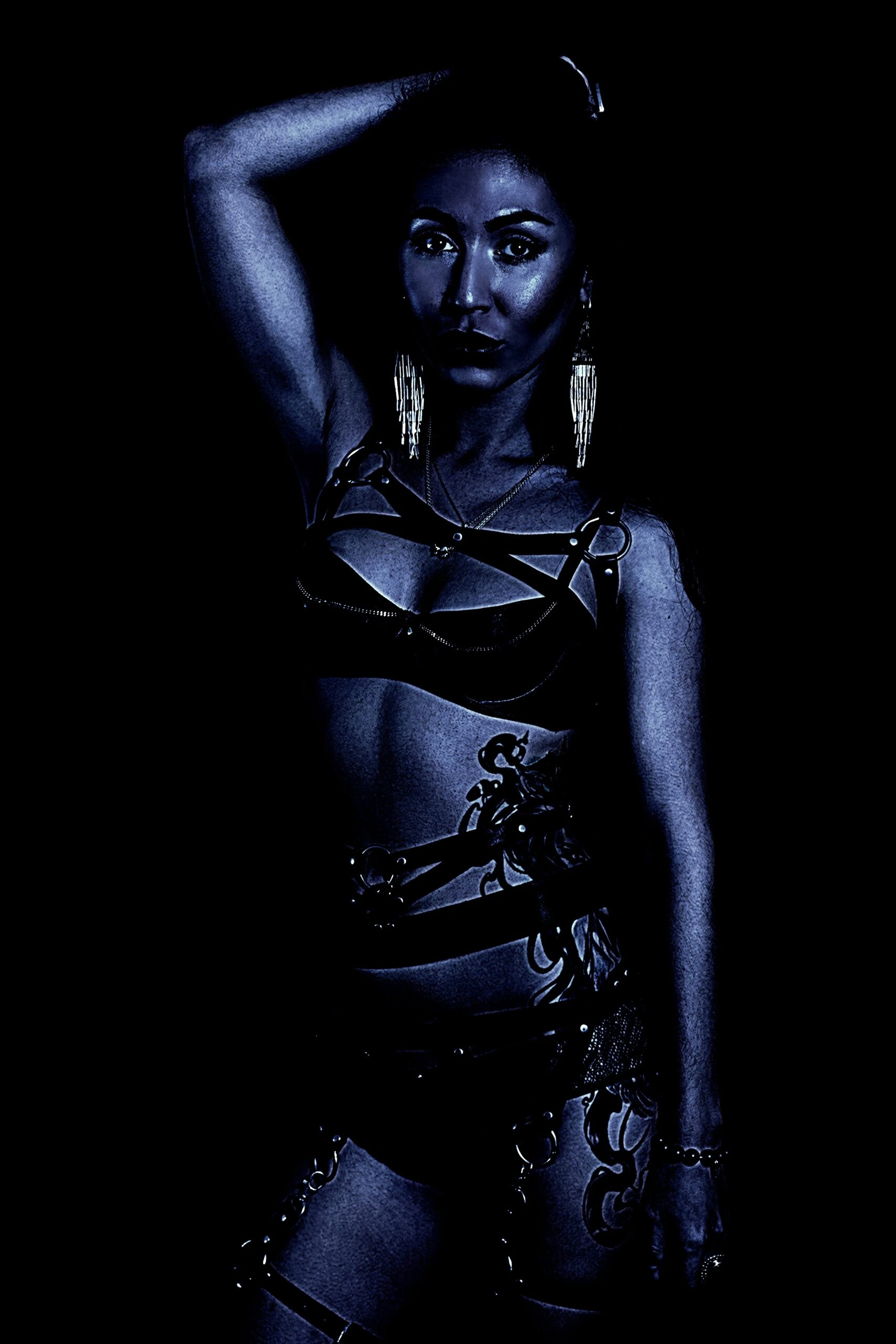 Woman in a harness and lingerie, posing with one arm raised, in a dark setting.