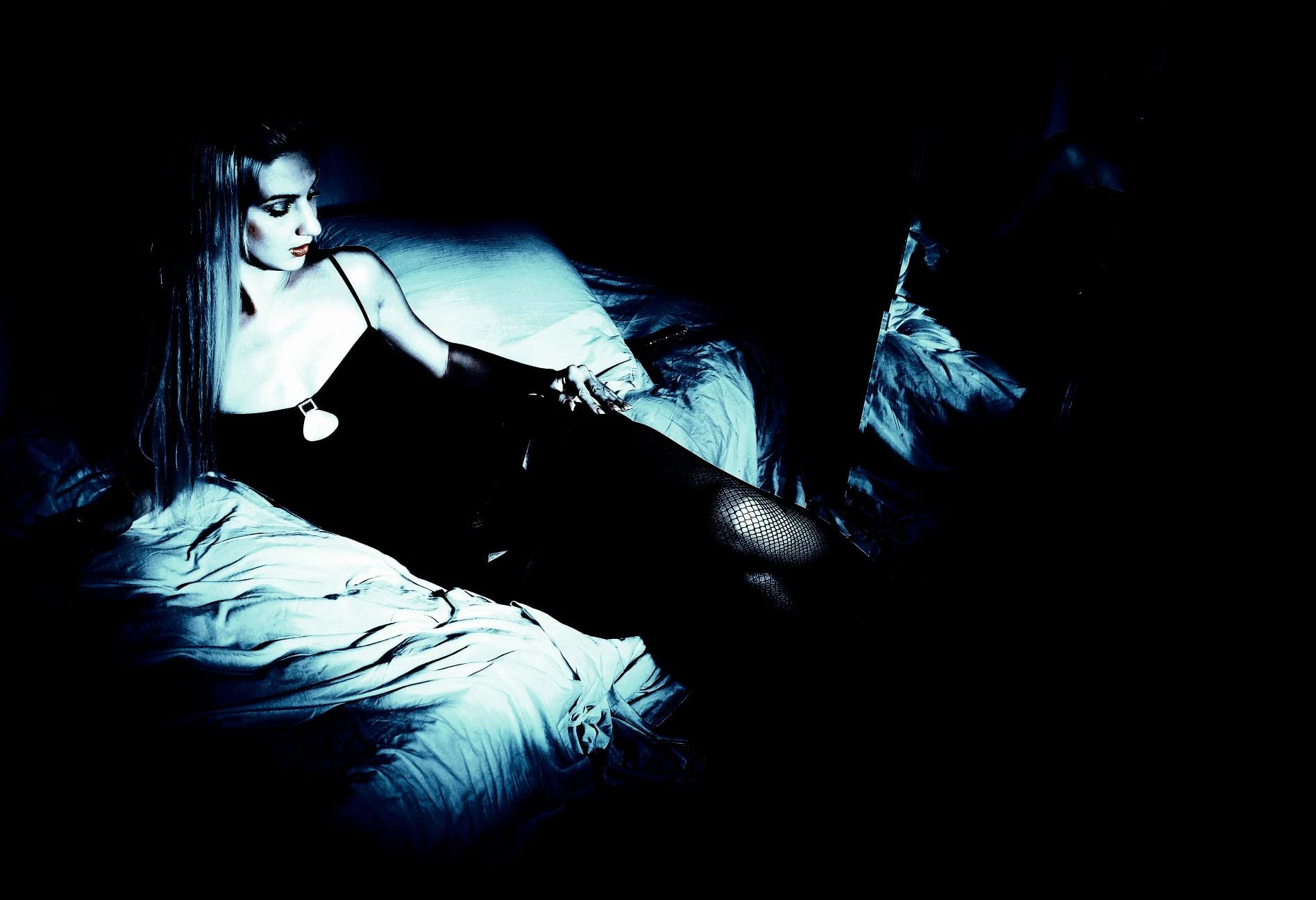 Woman in black dress reclining on a bed in low lighting, capturing a sultry, dark atmosphere.