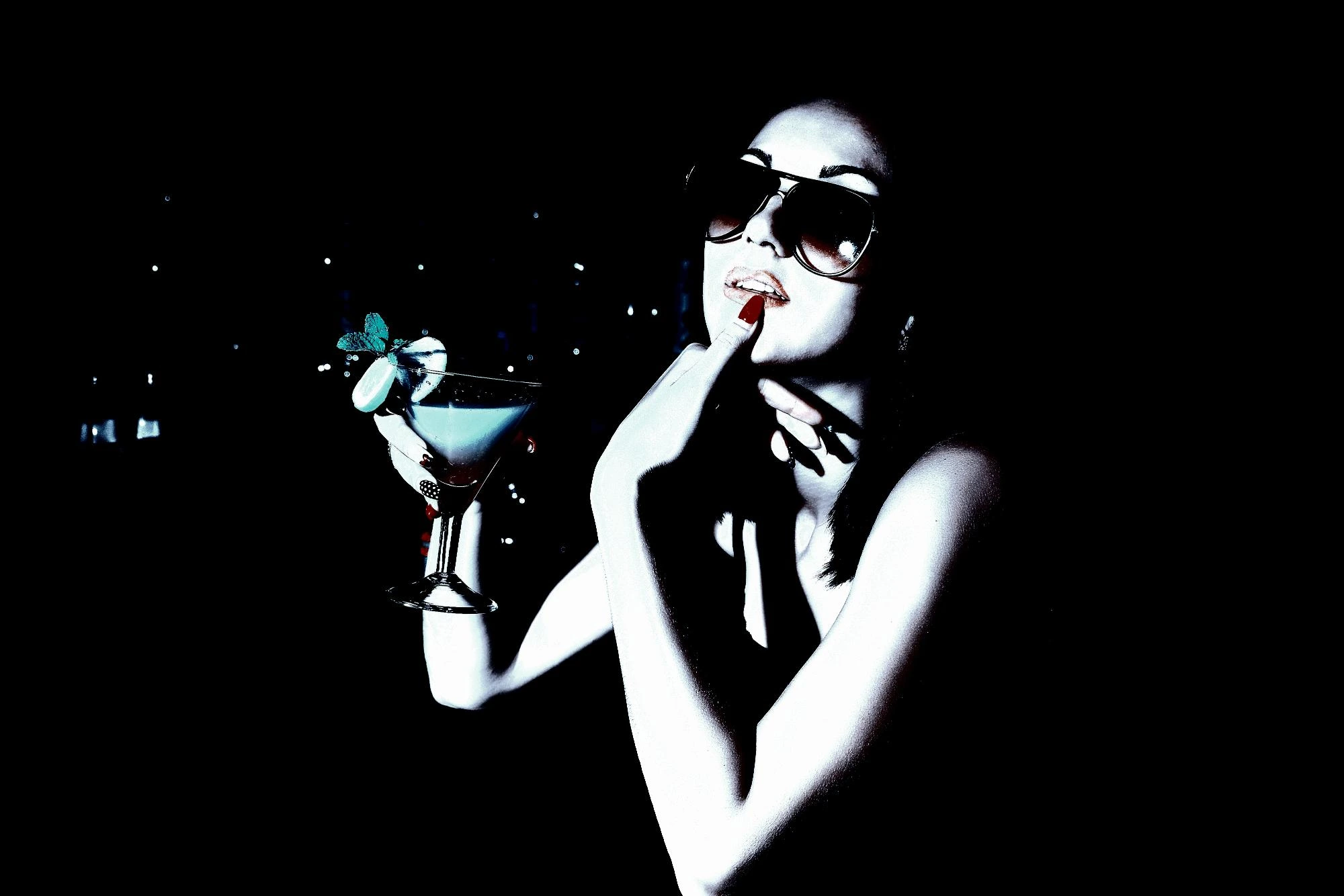 Woman with sunglasses and a cocktail, touching her lips, giving off a playful yet enigmatic vibe in low lighting.