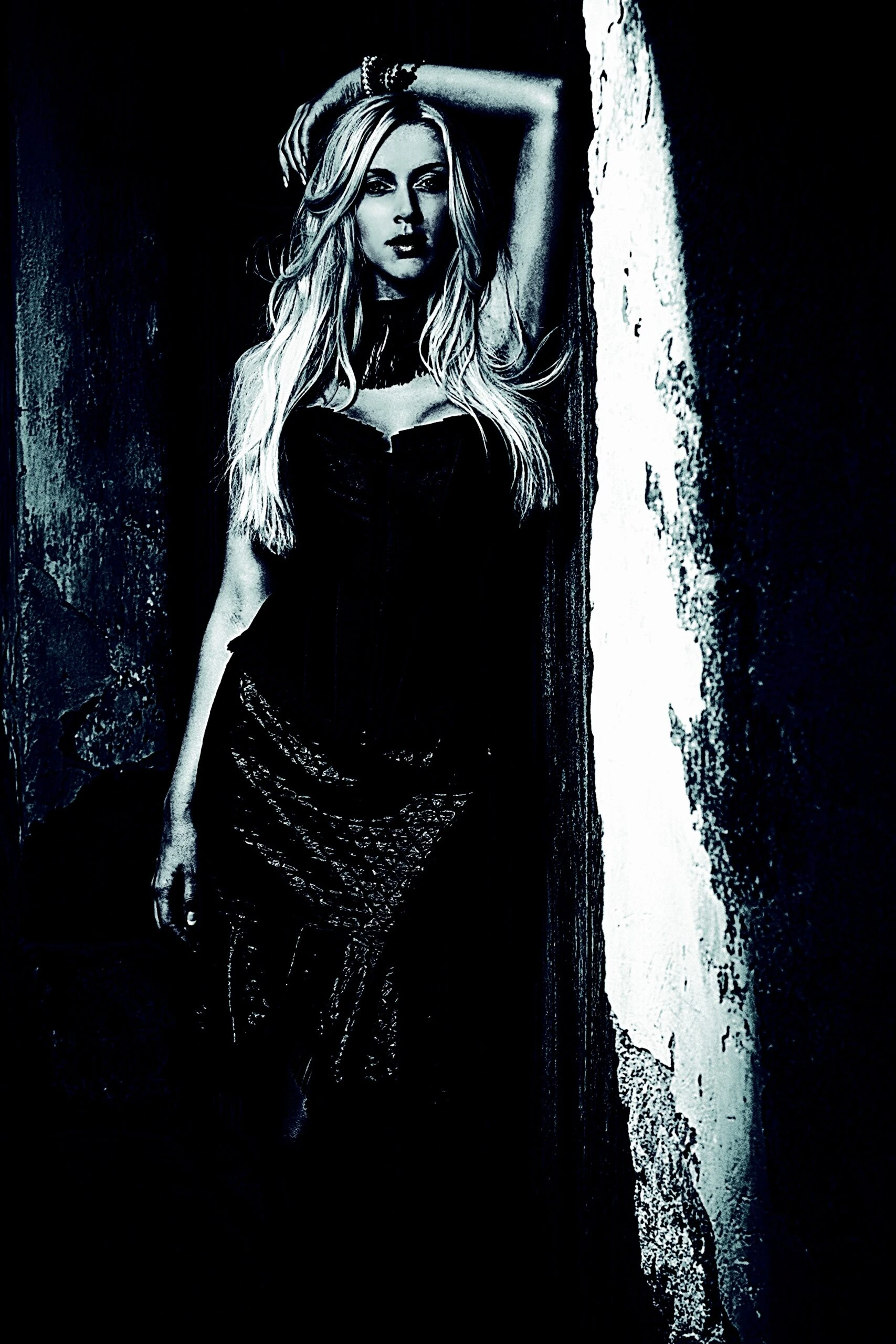 Blonde woman in a dark gothic outfit standing against a textured wall with a mysterious expression.