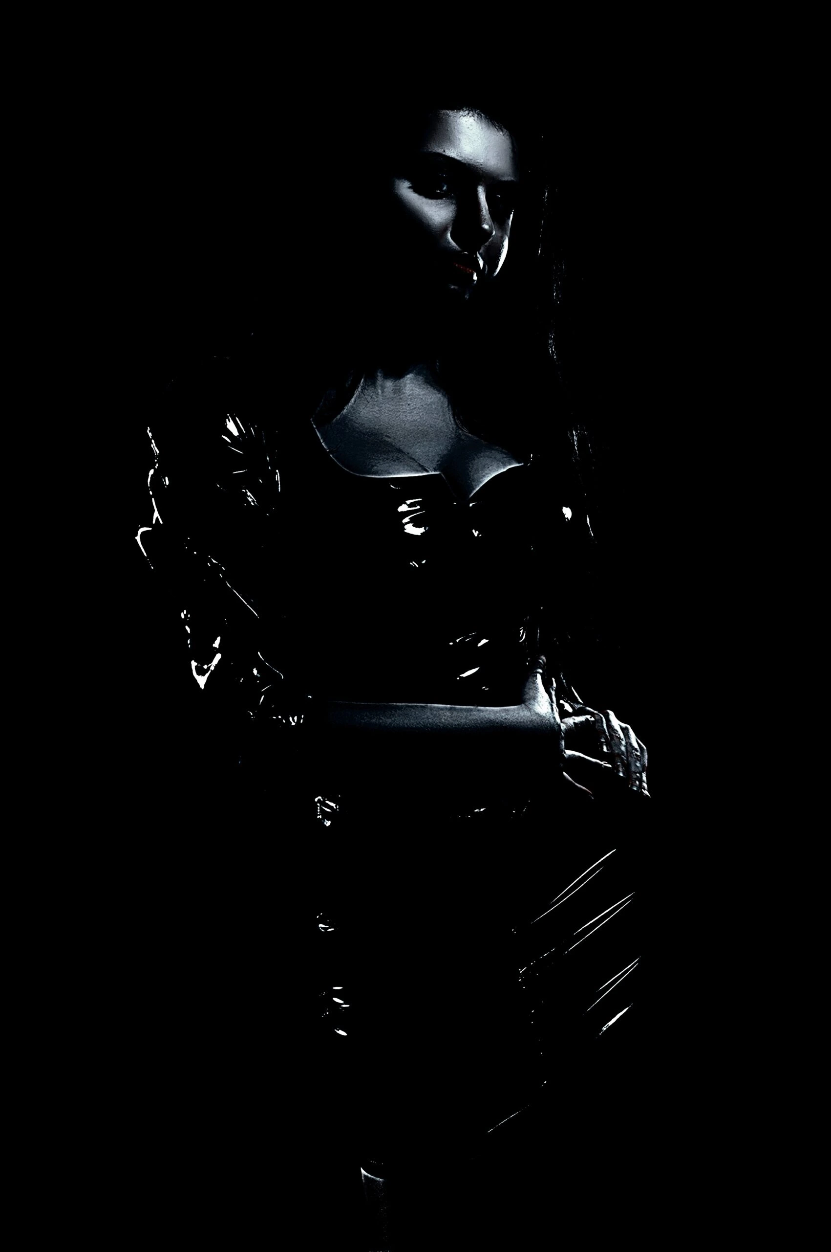 Woman in a shiny black latex outfit standing in the shadows with a dark, mysterious look.