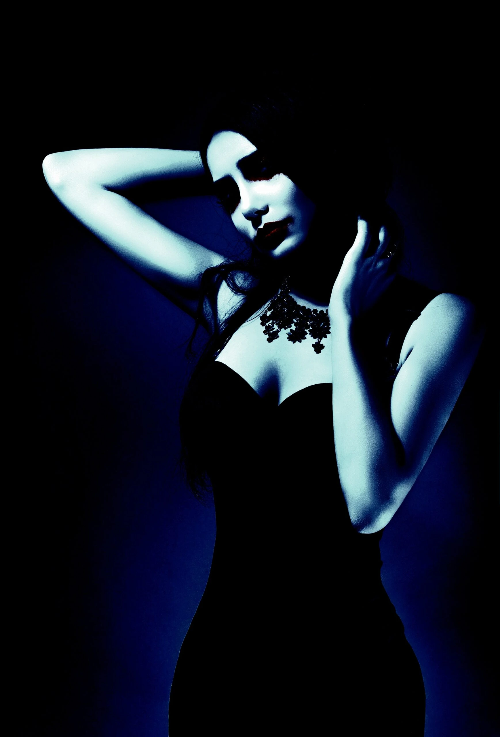 Woman in a black dress with a gothic necklace, posing with her hand on her head against a dark background.