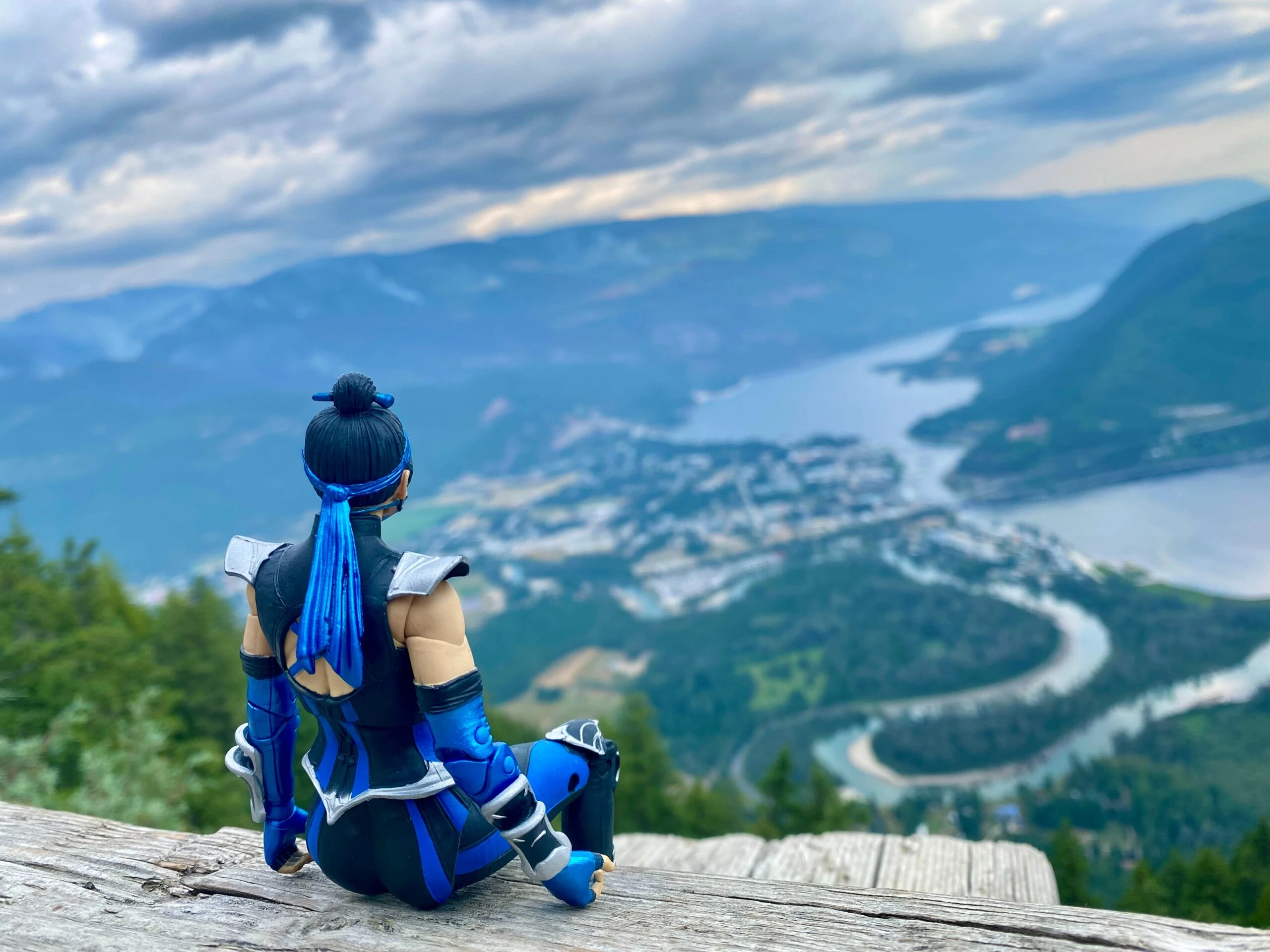 Figure of a warrior in blue armor sitting on a mountain ledge overlooking a scenic landscape.
