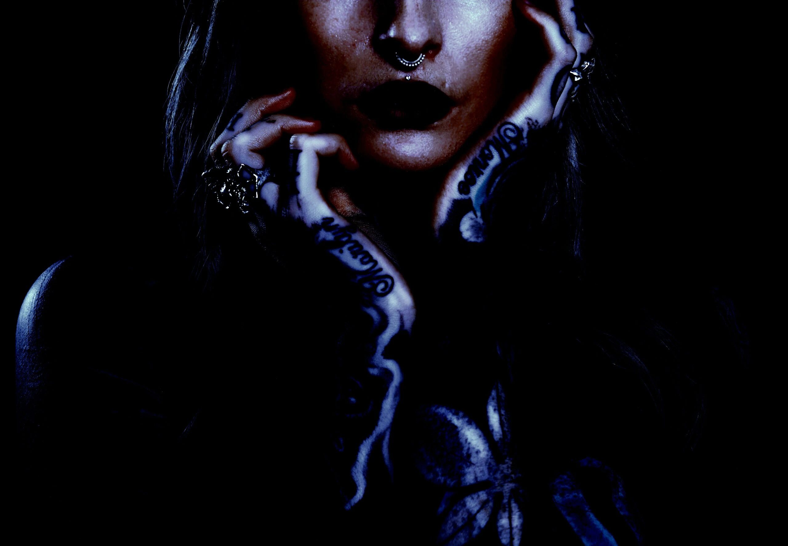 Close-up of a gothic woman with dark lipstick, septum piercing, and tattooed hands in a moody, shadowy setting.