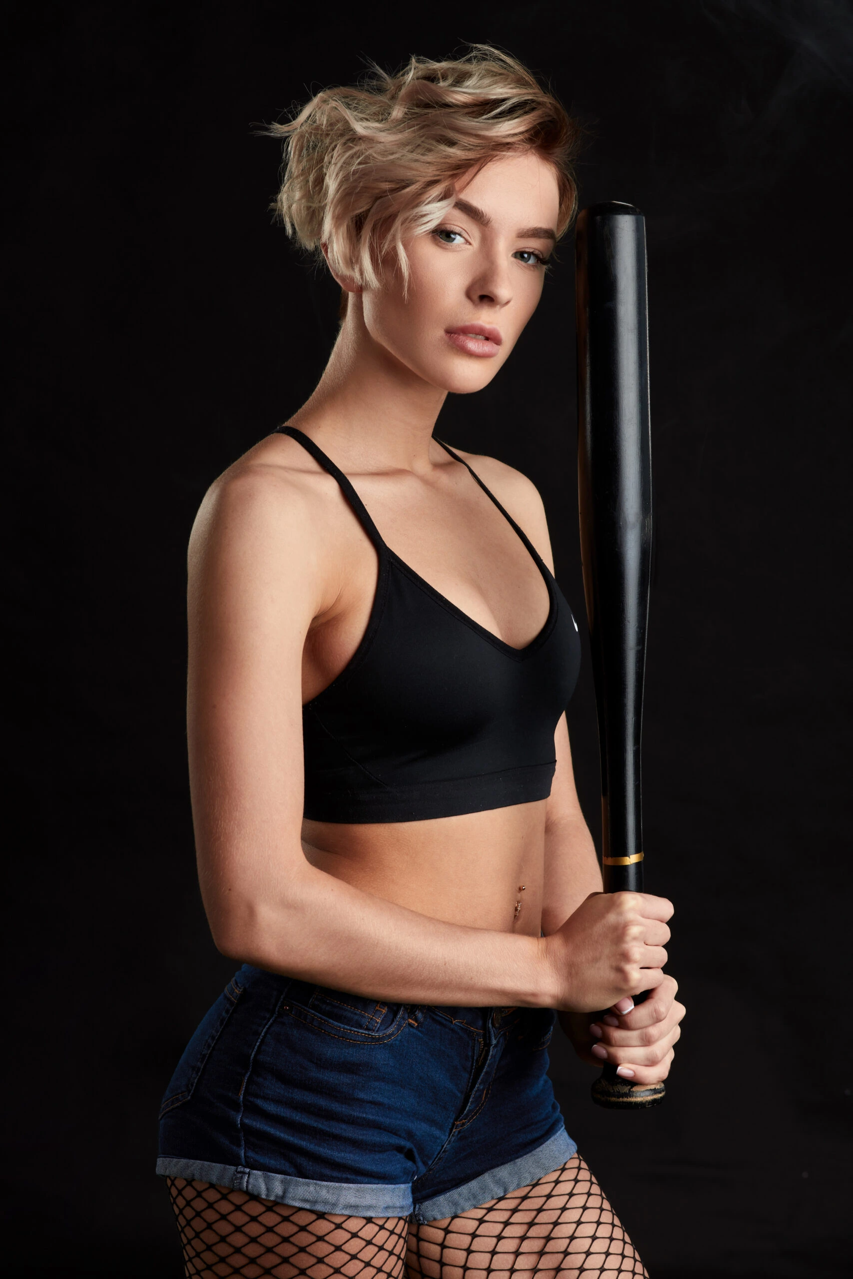 Woman with short blonde hair holding a baseball bat, wearing fishnet stockings and a black sports bra against a dark background.
