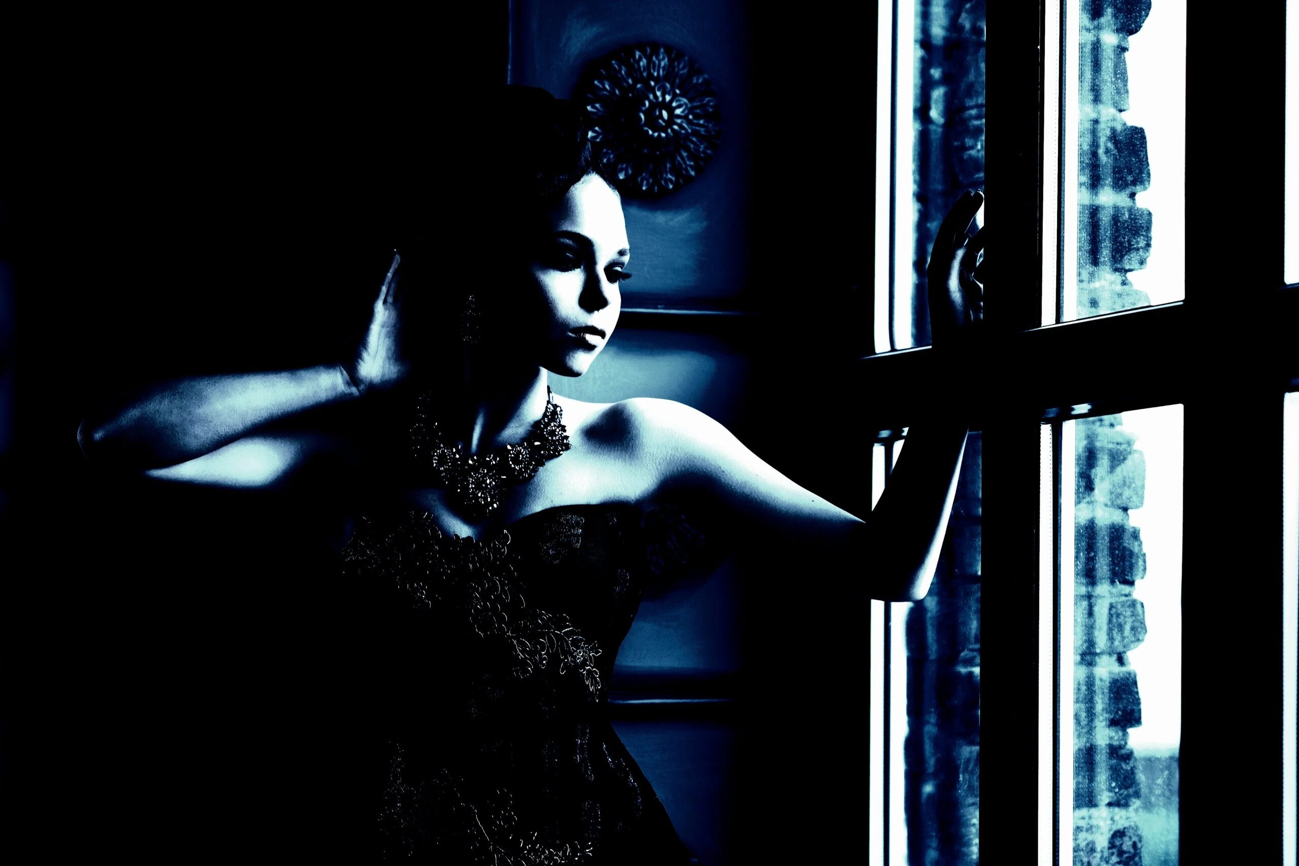 Woman in a gothic lace top and ornate necklace, gazing out a window with a contemplative expression in soft blue lighting.