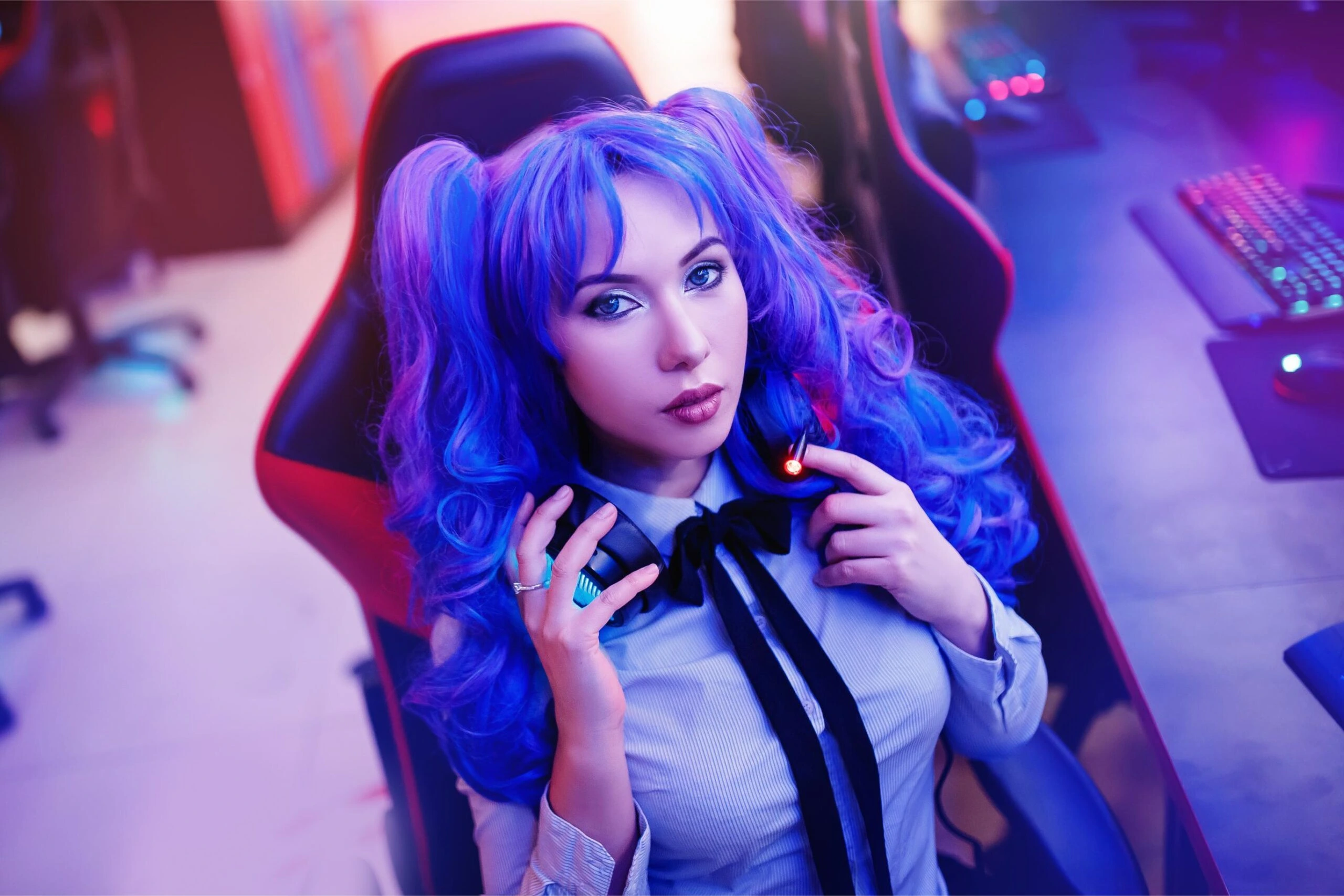 Woman with blue-purple hair in pigtails, seated in a gaming chair, holding headphones with a focused expression.