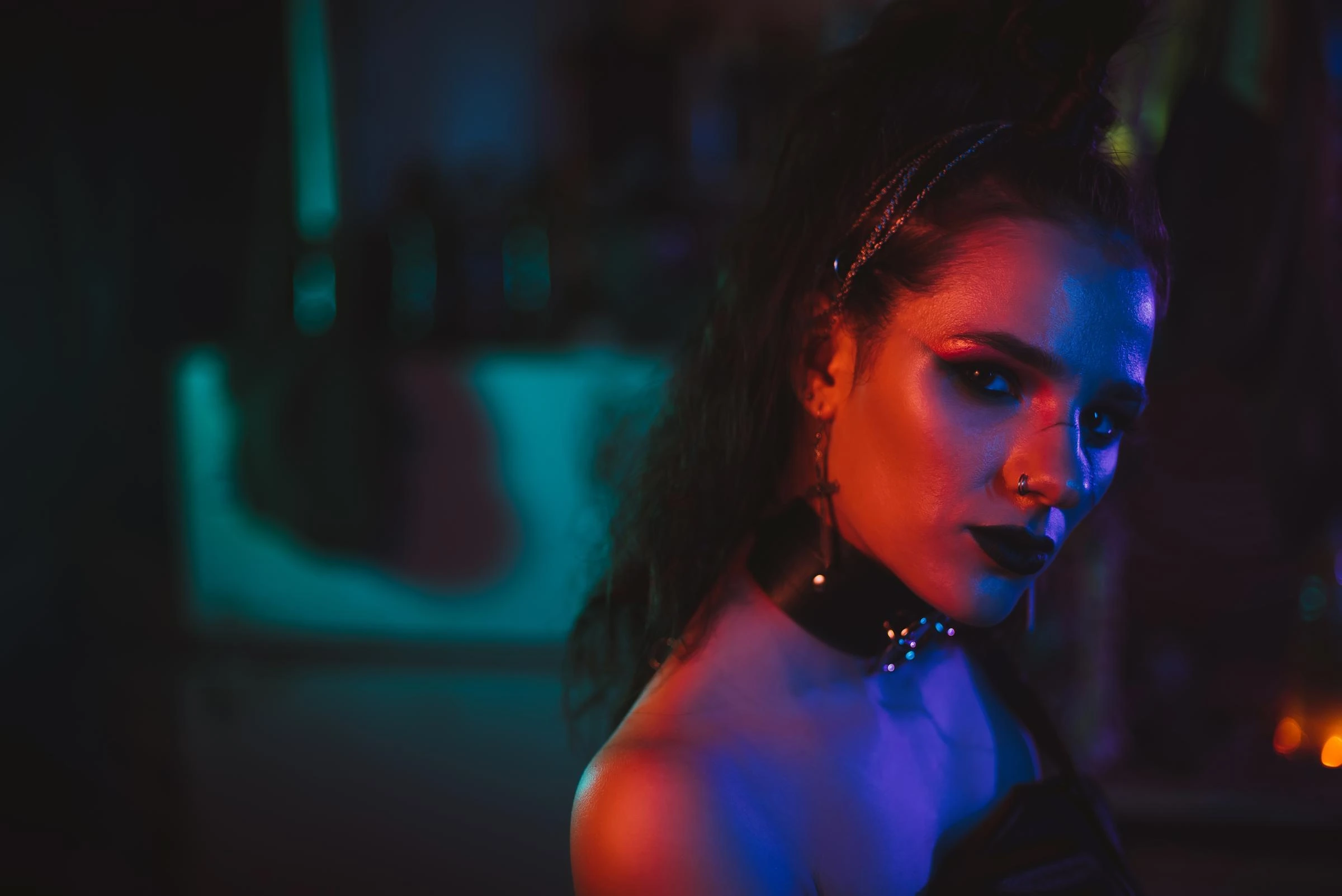 Gothic woman with dark makeup and piercing stare, wearing black choker, standing in a dimly lit room with neon lights.