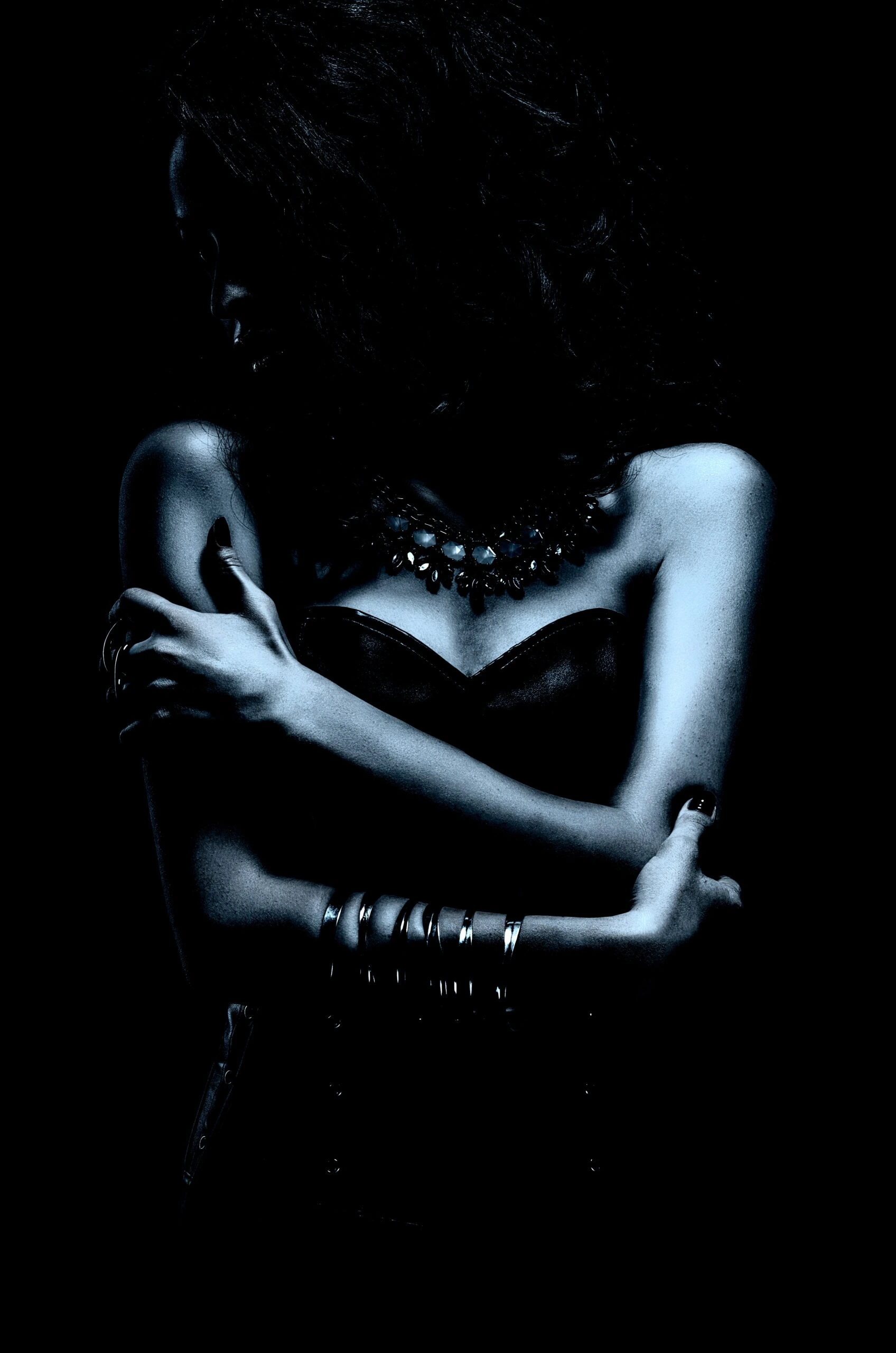 Mysterious woman in a strapless top, wearing a dark statement necklace, arms crossed against a black background.