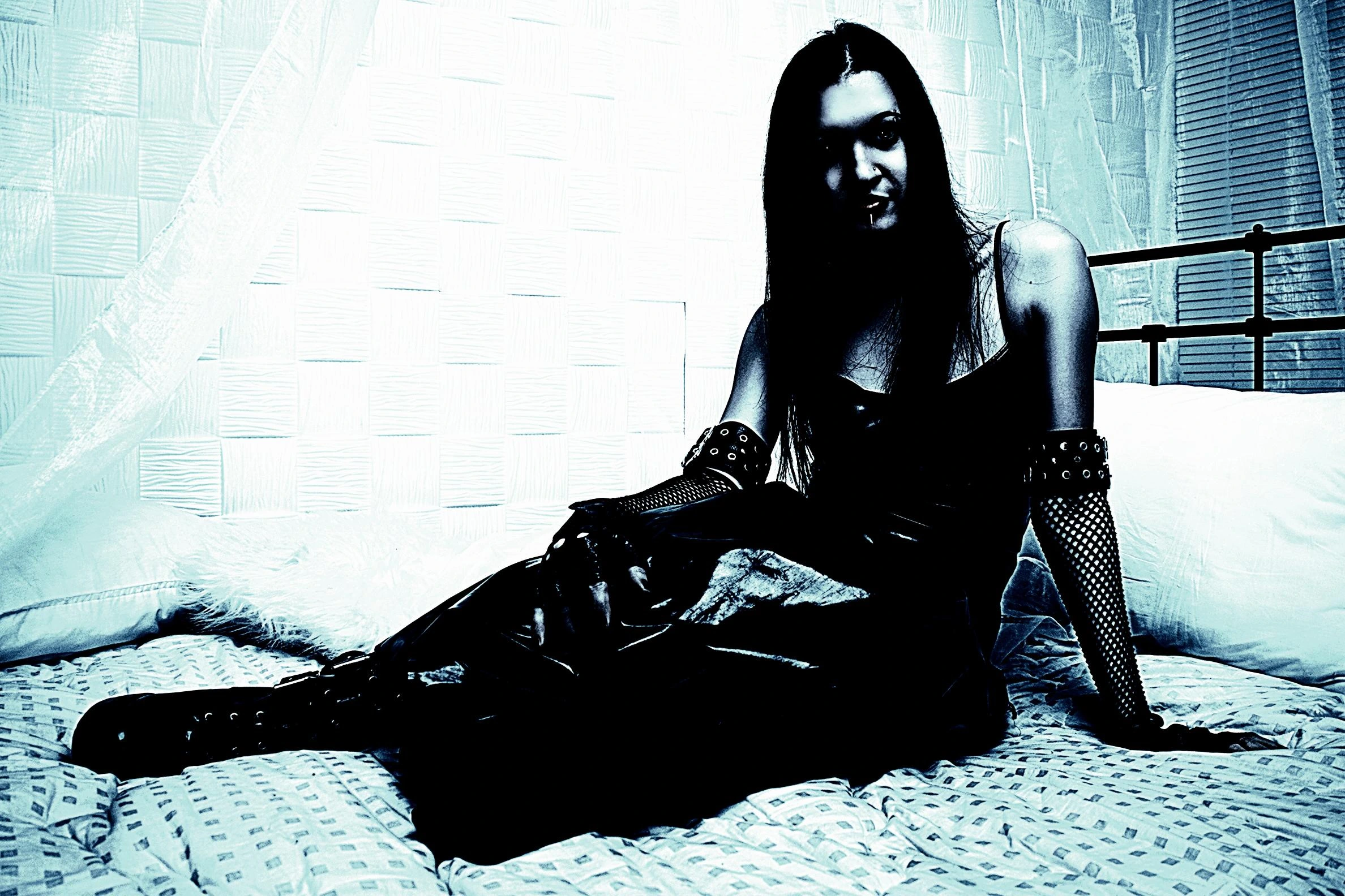 Woman sitting on a bed wearing a gothic black dress, with fishnet gloves and a mysterious expression.