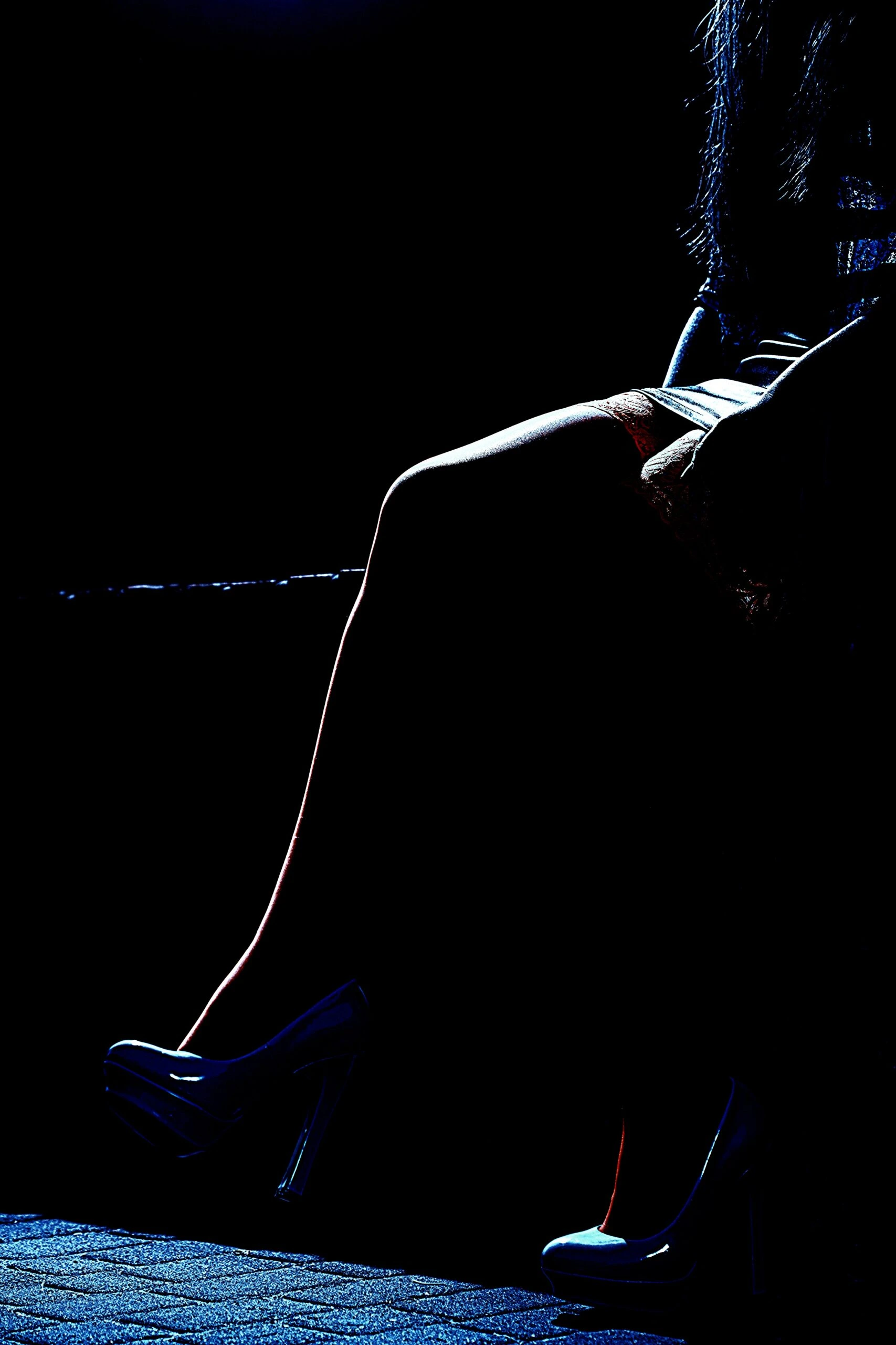 The legs of a woman in high heels, sitting in a dark, moody environment, with the focus on the curve of her leg and the reflective surface of her shoes.
