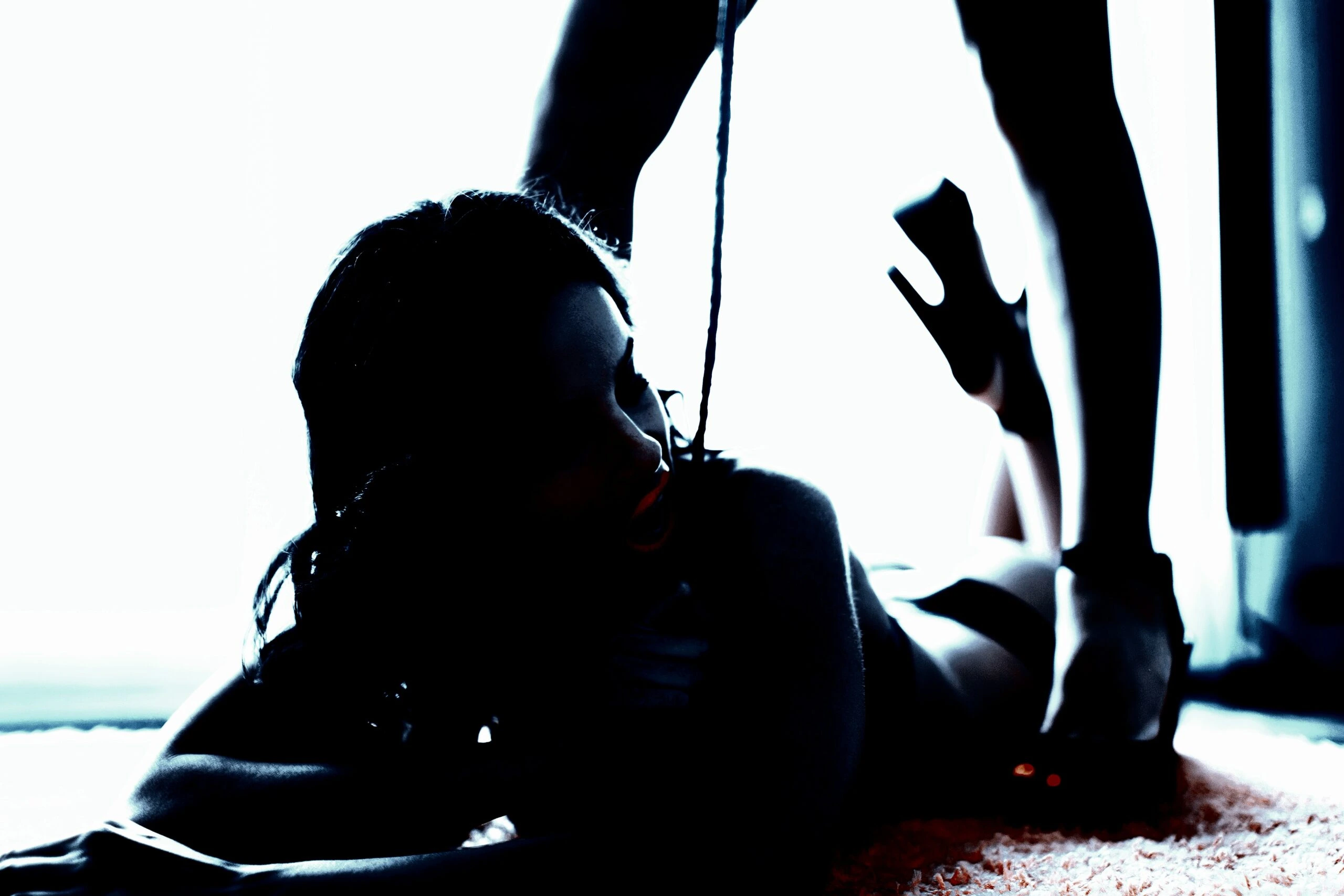 A dramatic, silhouette shot of a woman on the floor, submissive, while another person stands over her, their high heels highlighted in sharp contrast, creating a provocative and moody scene.