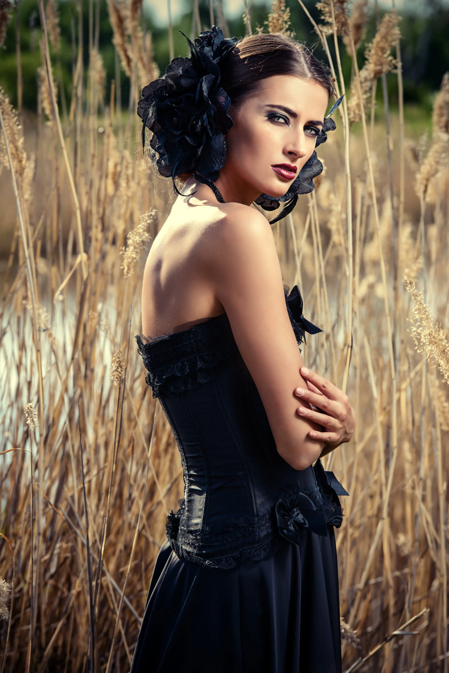 Elegant woman in a gothic-inspired black corset dress with dark floral headpiece, posing in a field of tall reeds.