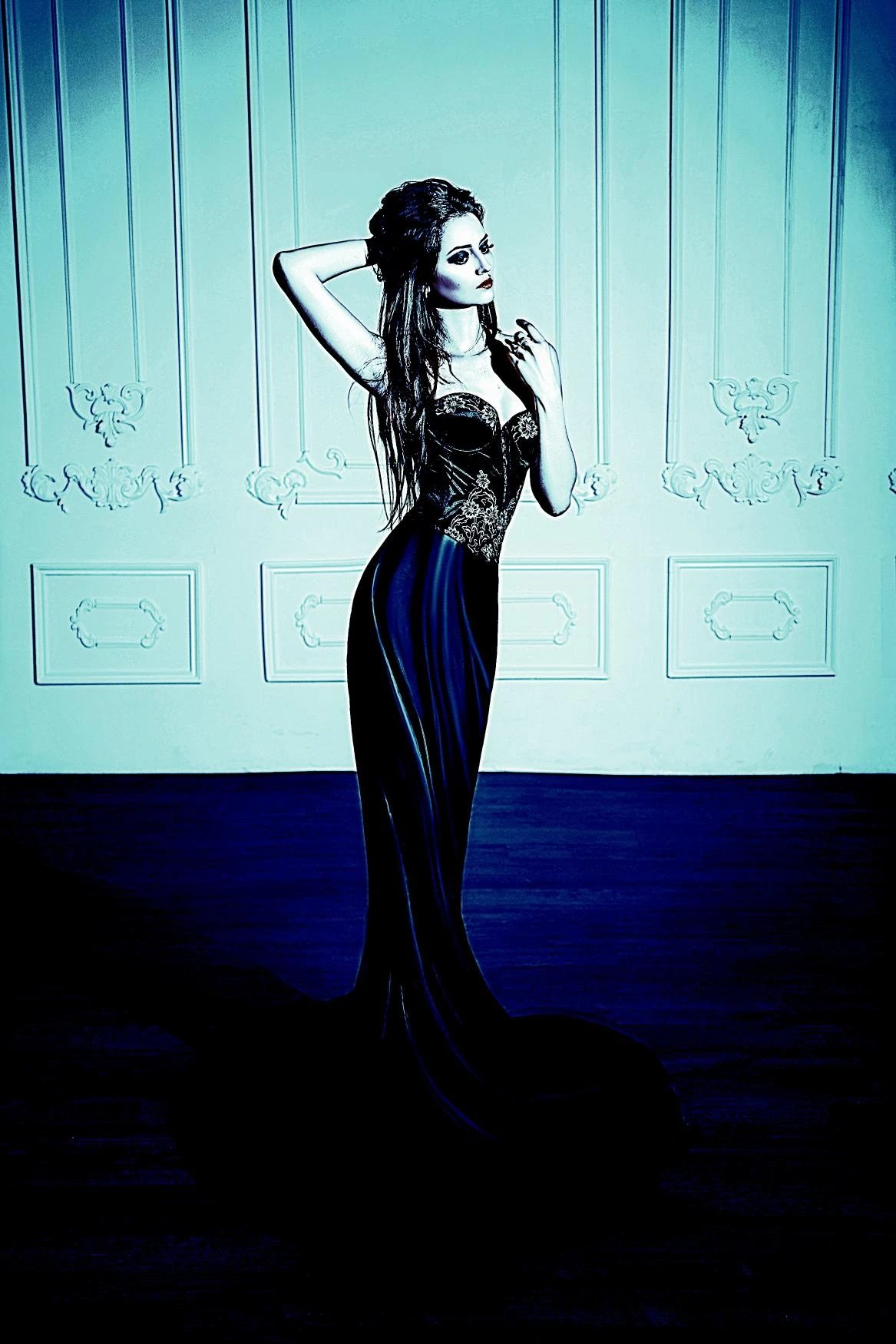 Model in a fitted black gothic dress with gold embroidery, posing against a sophisticated wall backdrop.