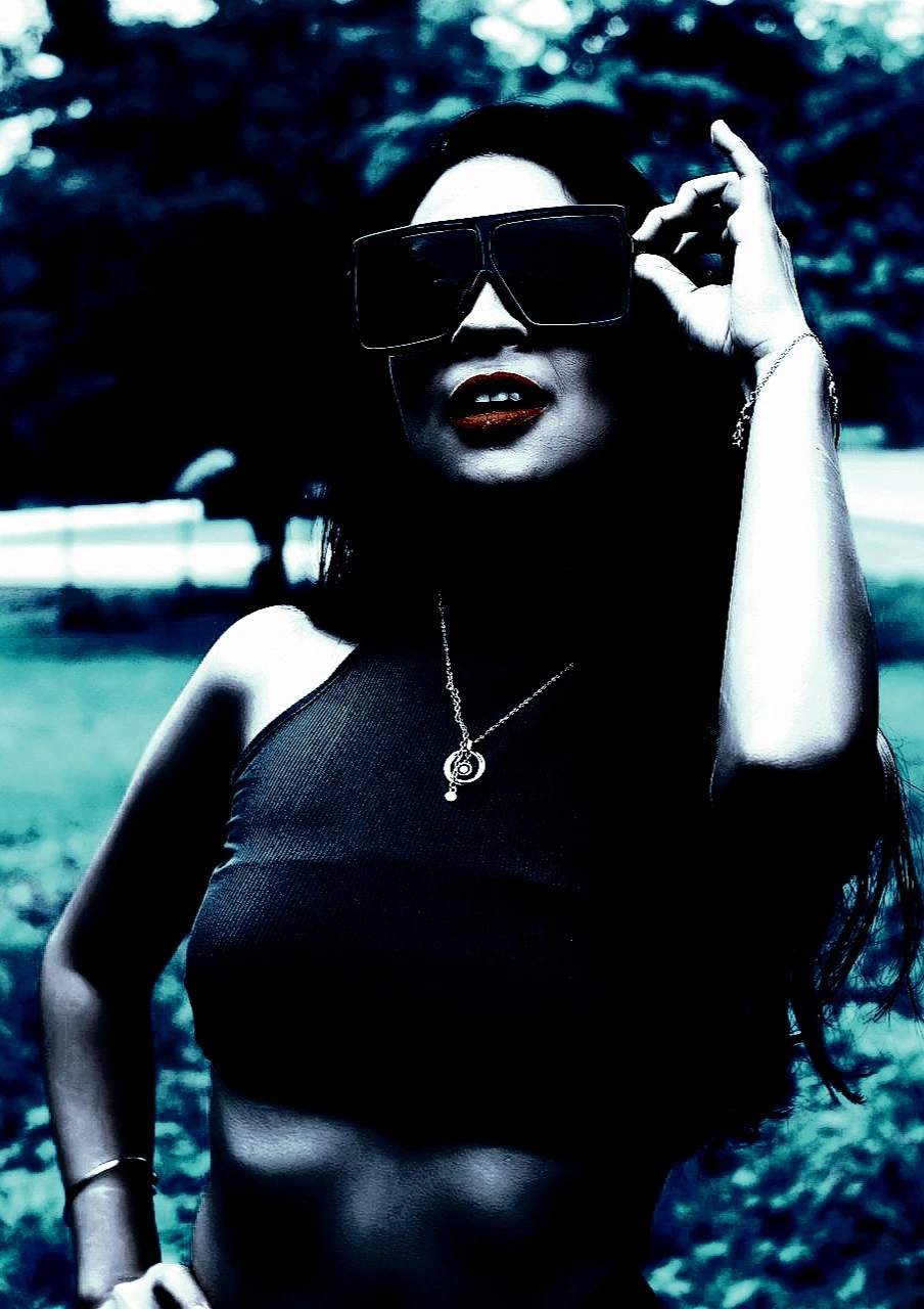 Woman wearing large black sunglasses with her hand on her hip, standing confidently against a green outdoor background.