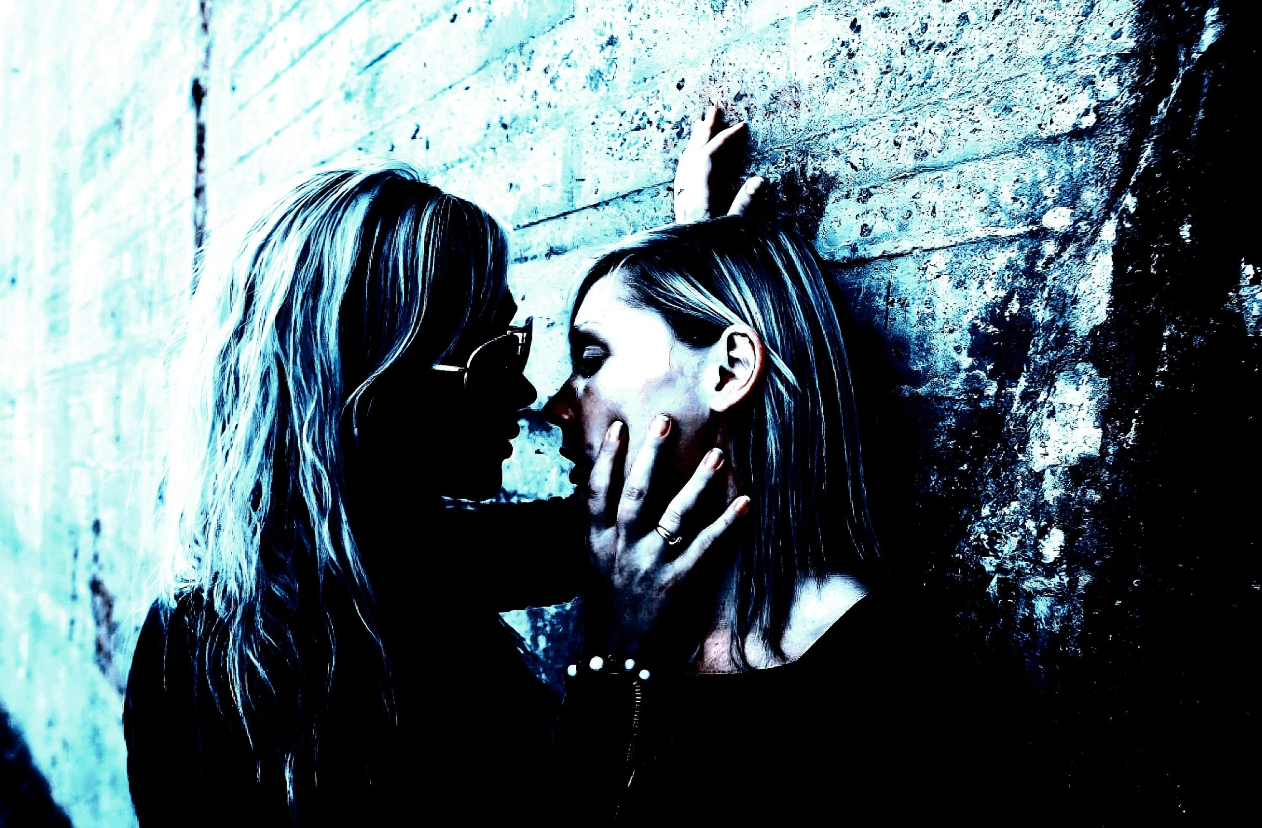 Two women in a close, intimate moment against a textured wall, bathed in a blue-toned light.