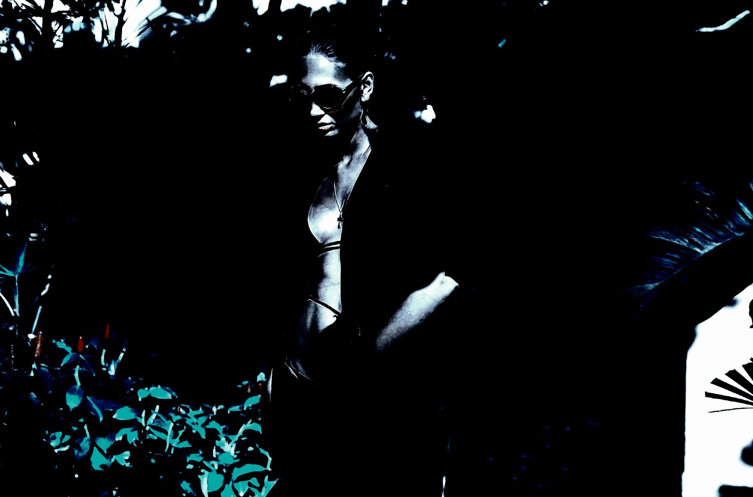 A woman standing outdoors, partially obscured by shadows, wearing dark sunglasses and a revealing top, blending into the deep shadows of the surrounding greenery. 