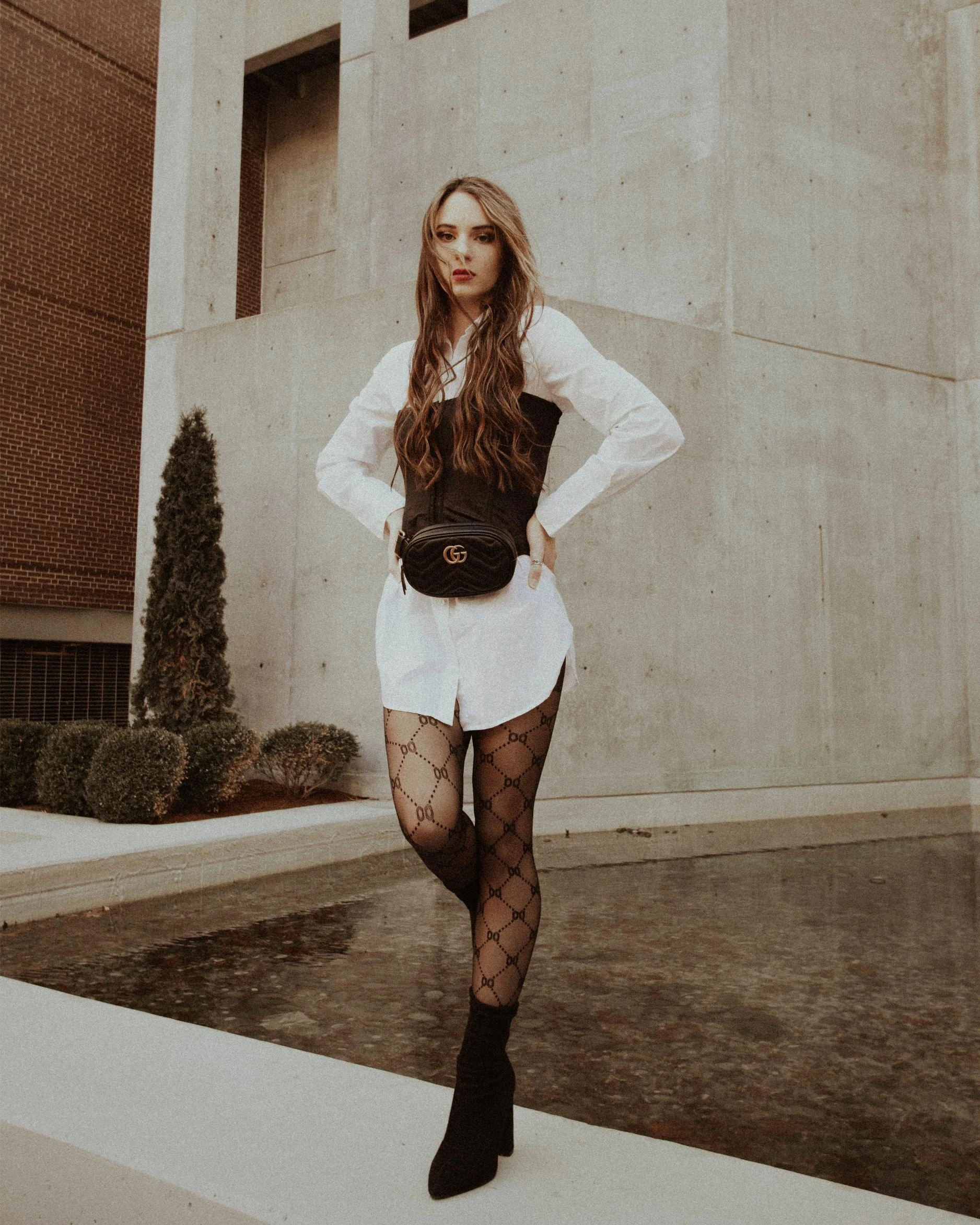 Stylish woman in black underbust corset over a white shirt dress, accessorized with a Gucci belt bag, standing against a modern concrete backdrop.