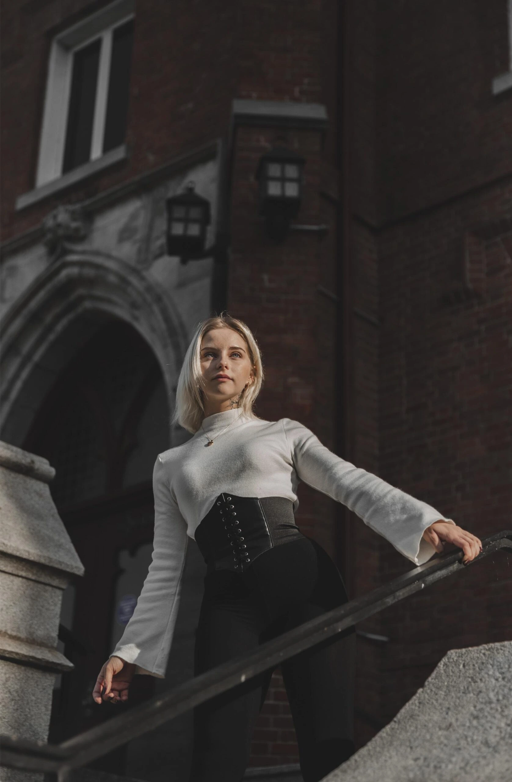 Blonde woman wearing a black underbust corset over a white sweater, standing on stone steps of a historic building.