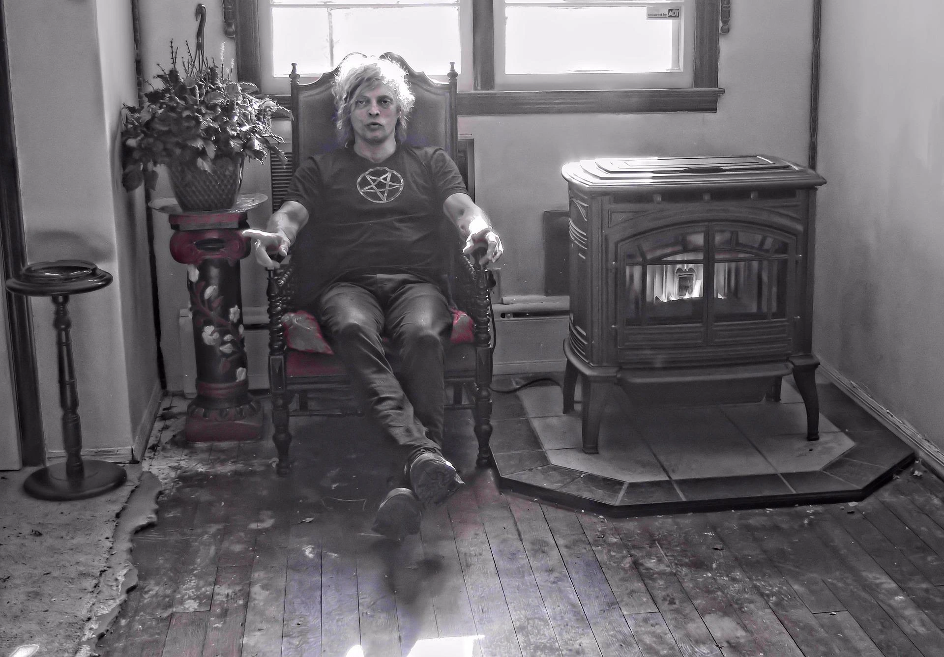 Man wearing a pentagram shirt, sitting in an ornate chair next to a vintage fireplace, in a shadowy room.
