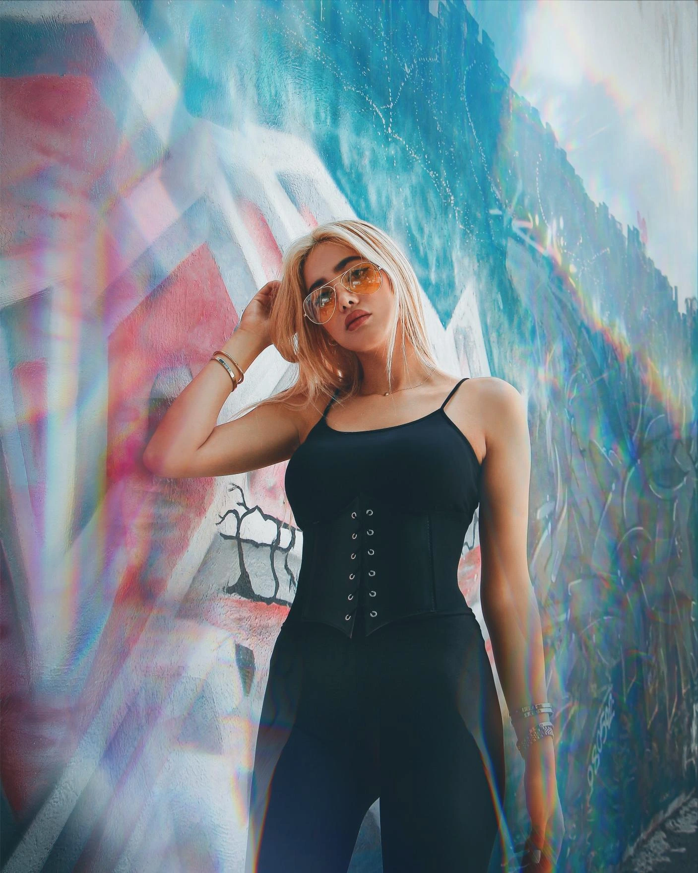 Blonde woman in black underbust corset and black leggings posing against a graffiti-covered wall with a colorful light prism effect.