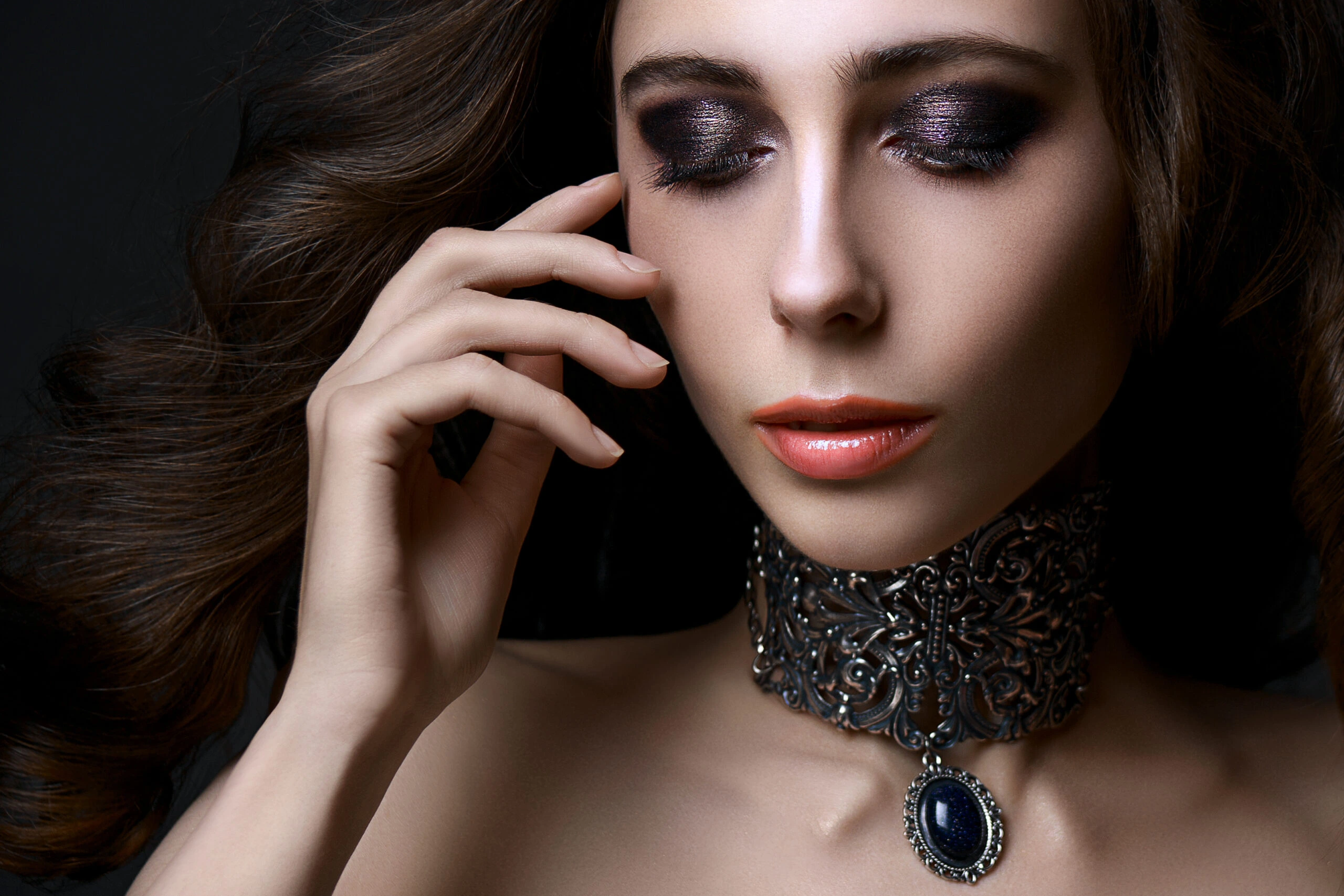 Woman with long flowing hair and dark eye makeup, wearing an ornate silver choker with a dark gemstone pendant.