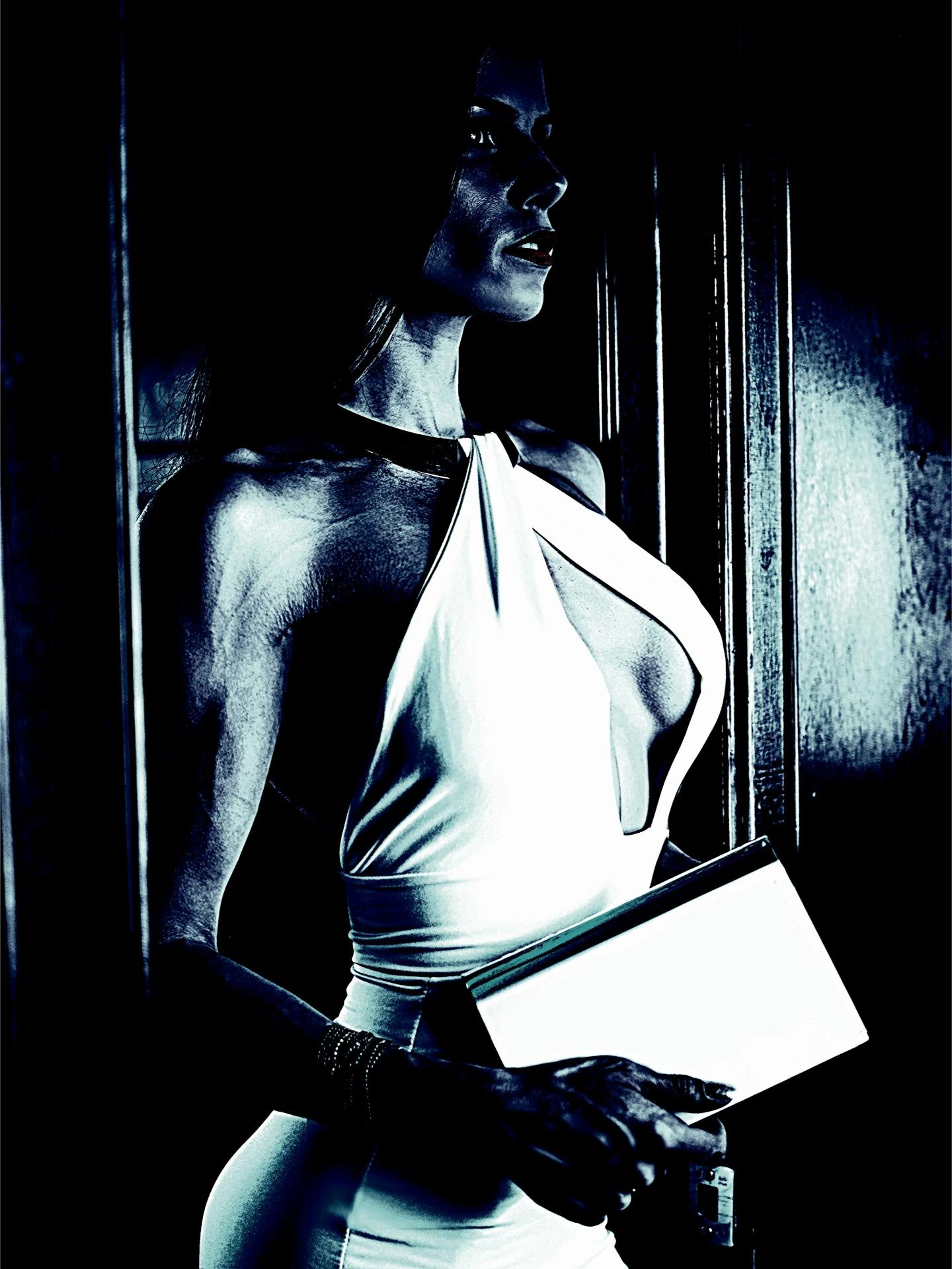 A woman in a sleek white halter dress holding a book, highlighted by shadows in a moody, gothic setting.