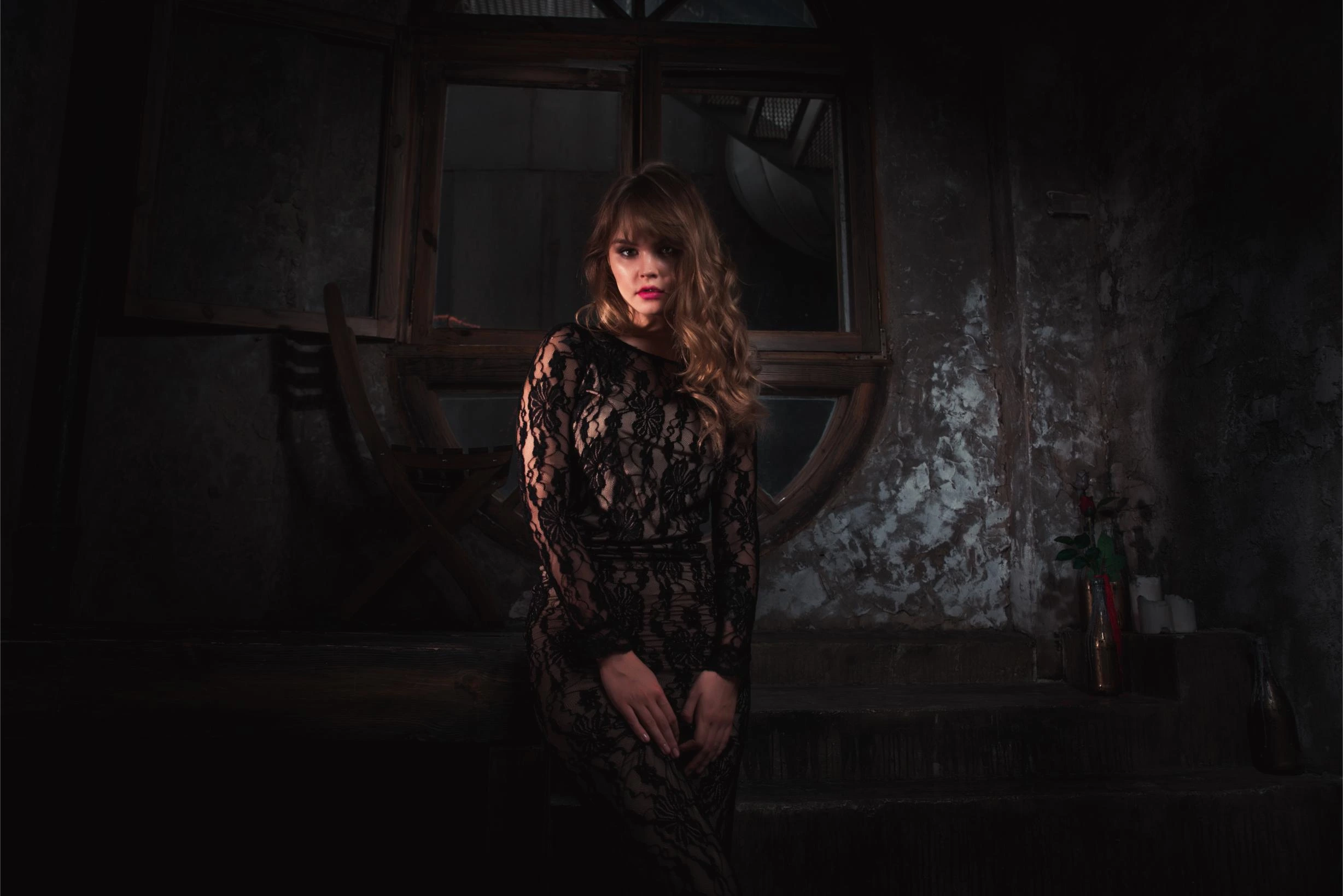 Woman in a lace-covered gothic black dress sitting in a moody, rustic interior, illuminated by dim lighting.