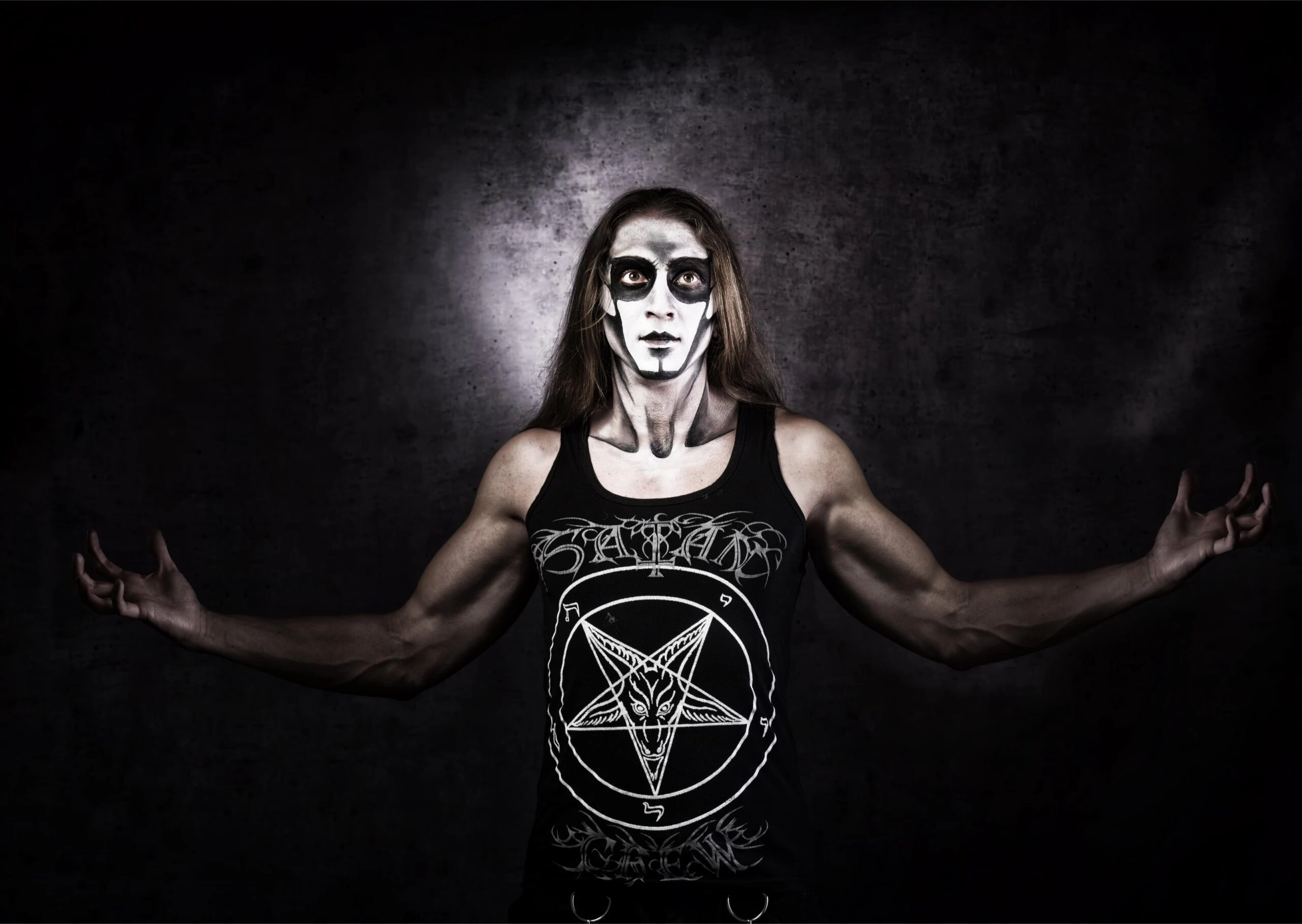 A man in corpse paint and a pentagram tank top, standing with arms outstretched in a dark and ominous setting.