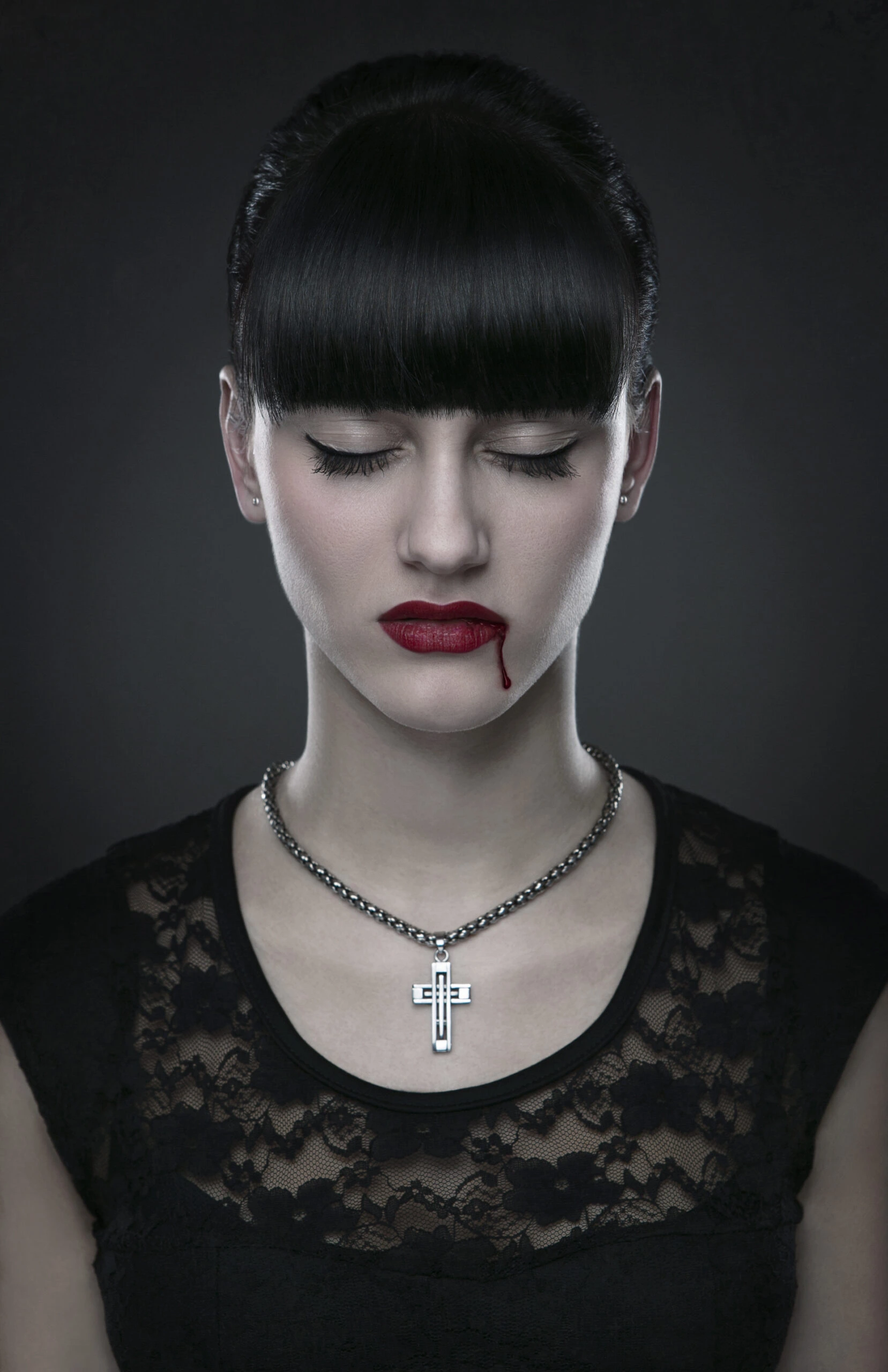 Woman with black hair and red lips, wearing a cross necklace, with a small drop of blood on her lip.