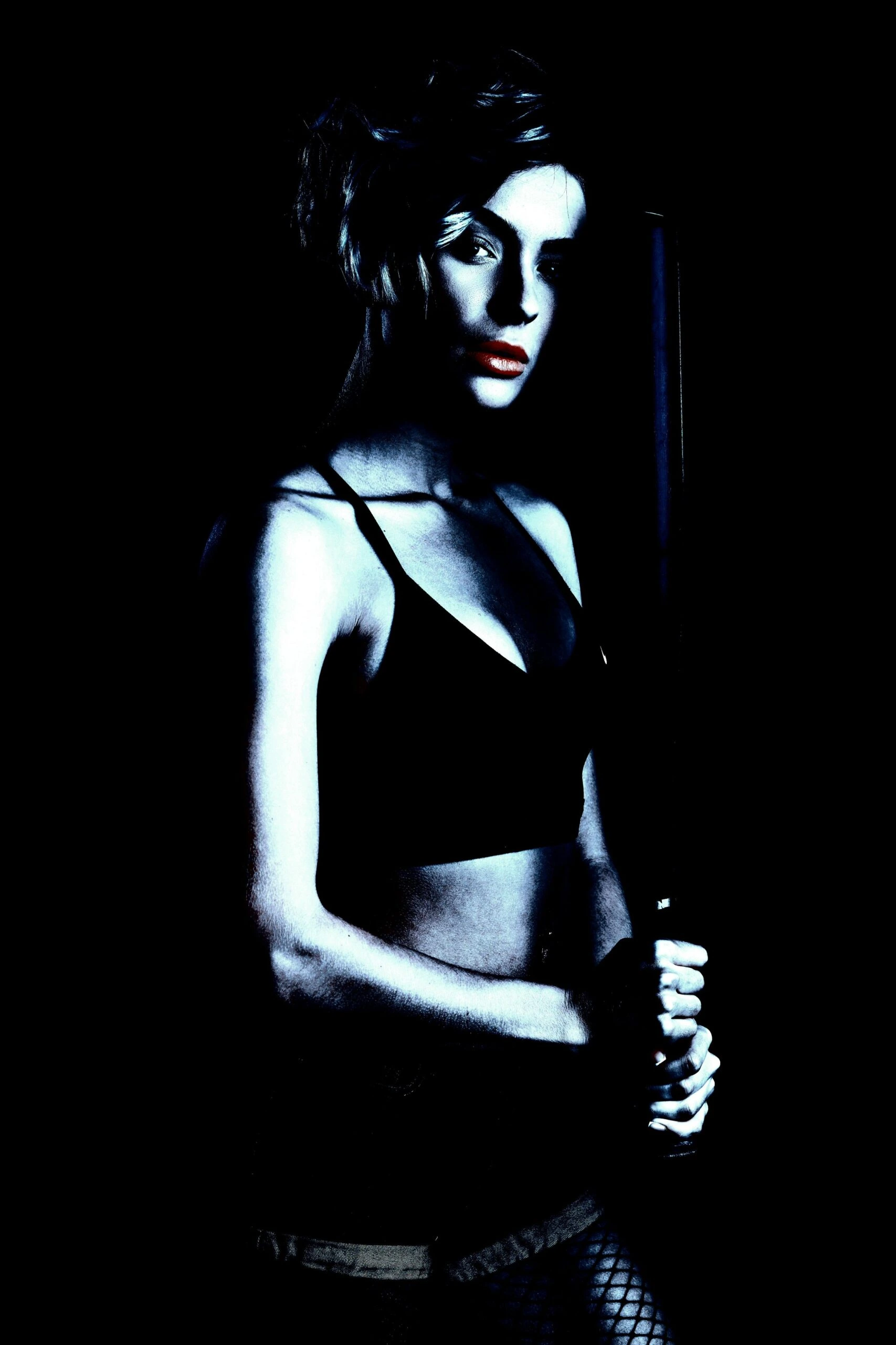 Dark, mysterious woman holding a baseball bat, dressed in athletic wear and fishnet stockings against a black backdrop.
