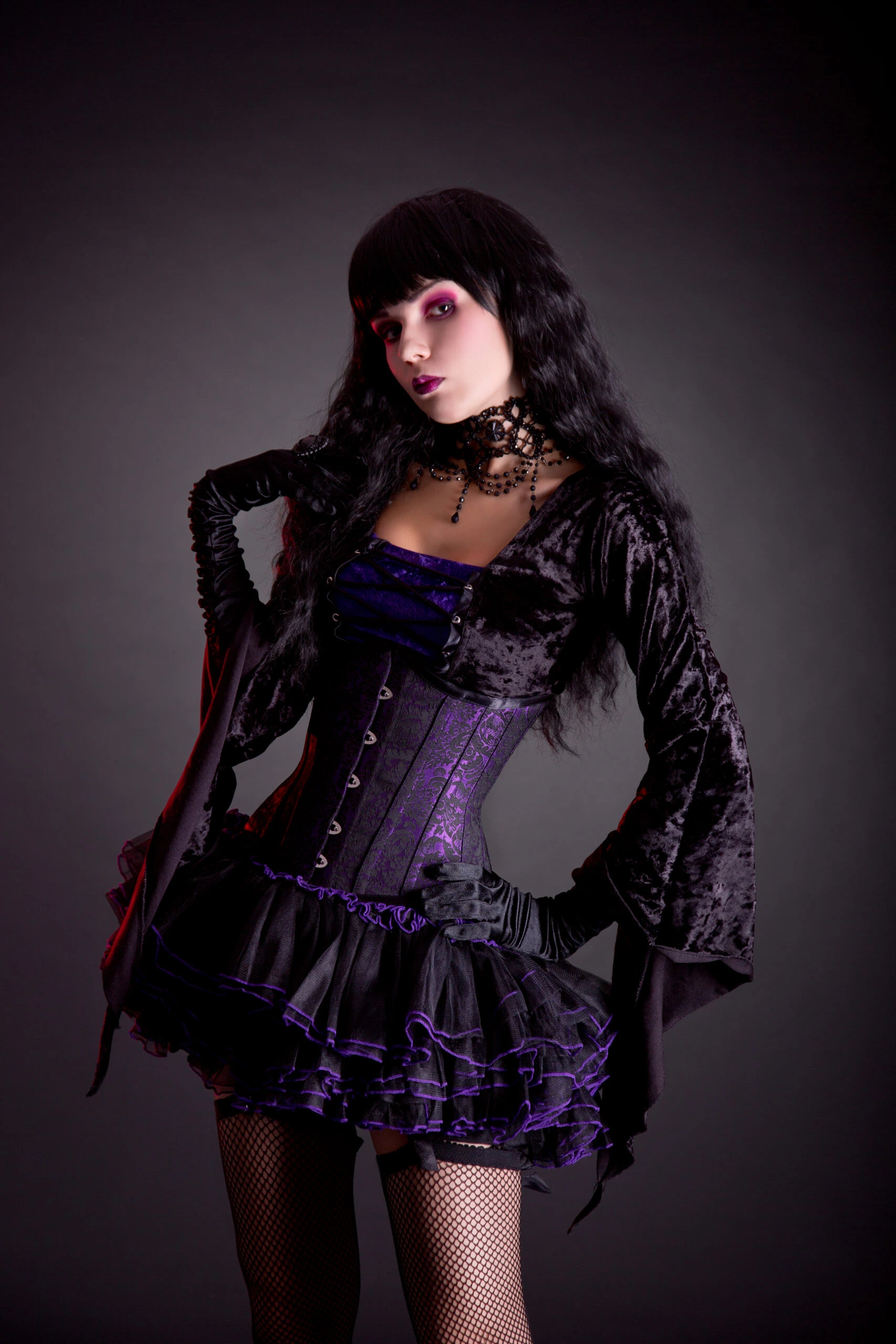 Woman dressed in a gothic outfit with a purple corset and black lace, posing confidently against a dark backdrop.