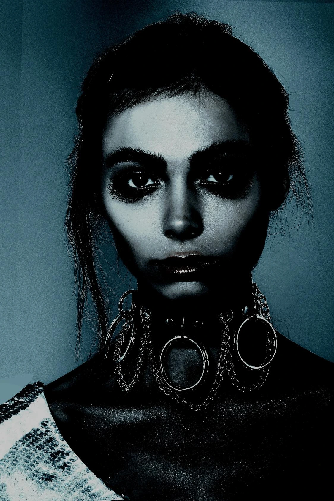A woman in a high-contrast image wearing a gothic chain choker with large metallic rings, set against a dark blue background.
