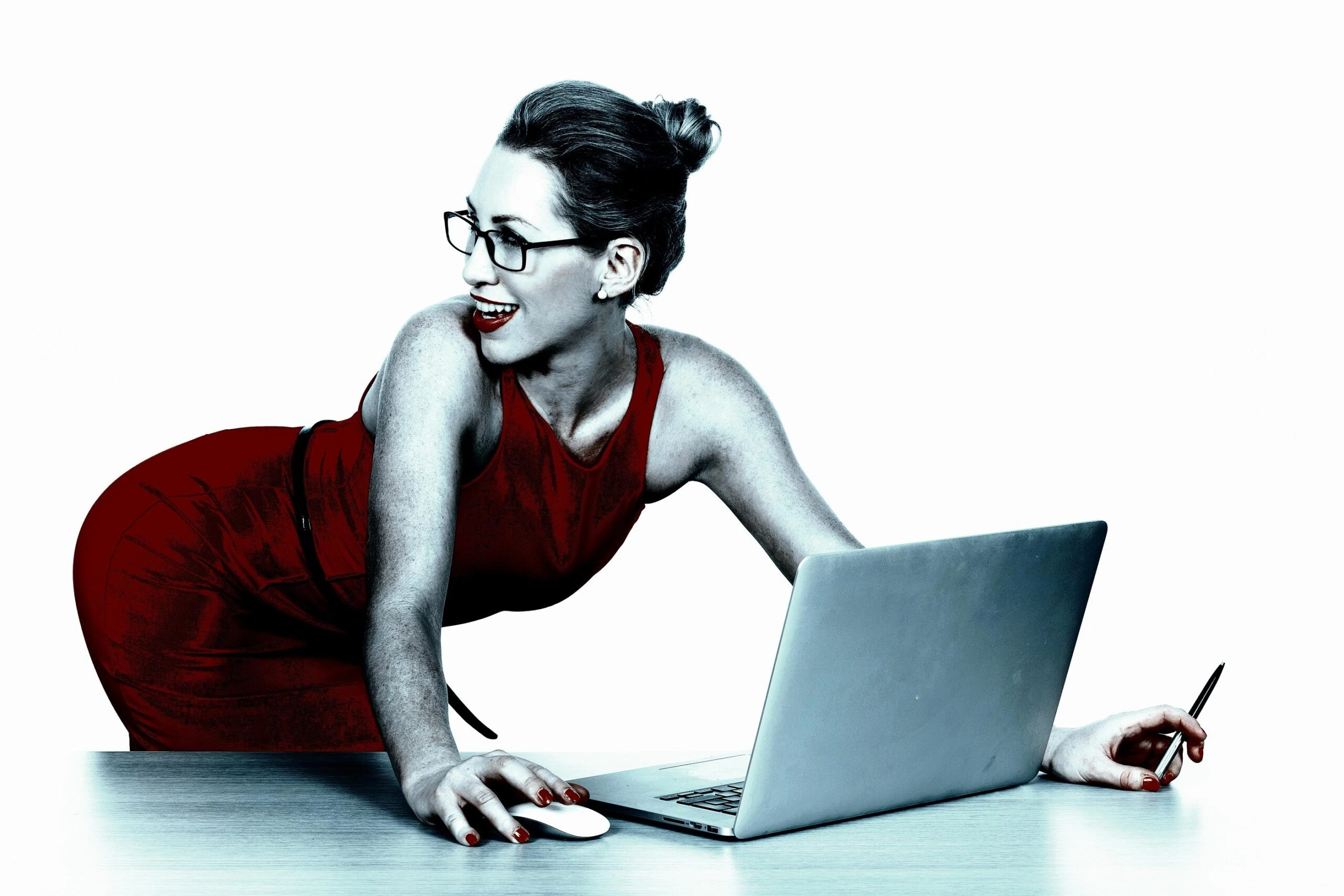 Businesswoman in a red dress leaning over a desk, smiling confidently while working on a laptop.
