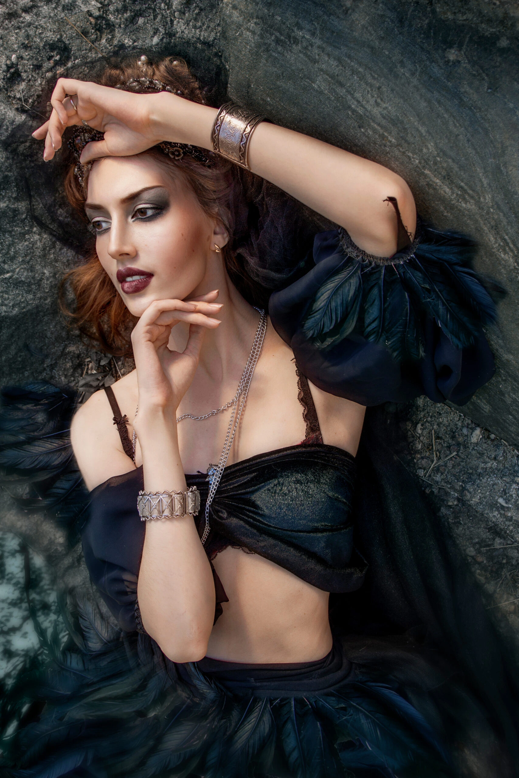 Mystical woman with dark makeup lying on a rock, adorned with silver jewelry and a black feathered outfit.