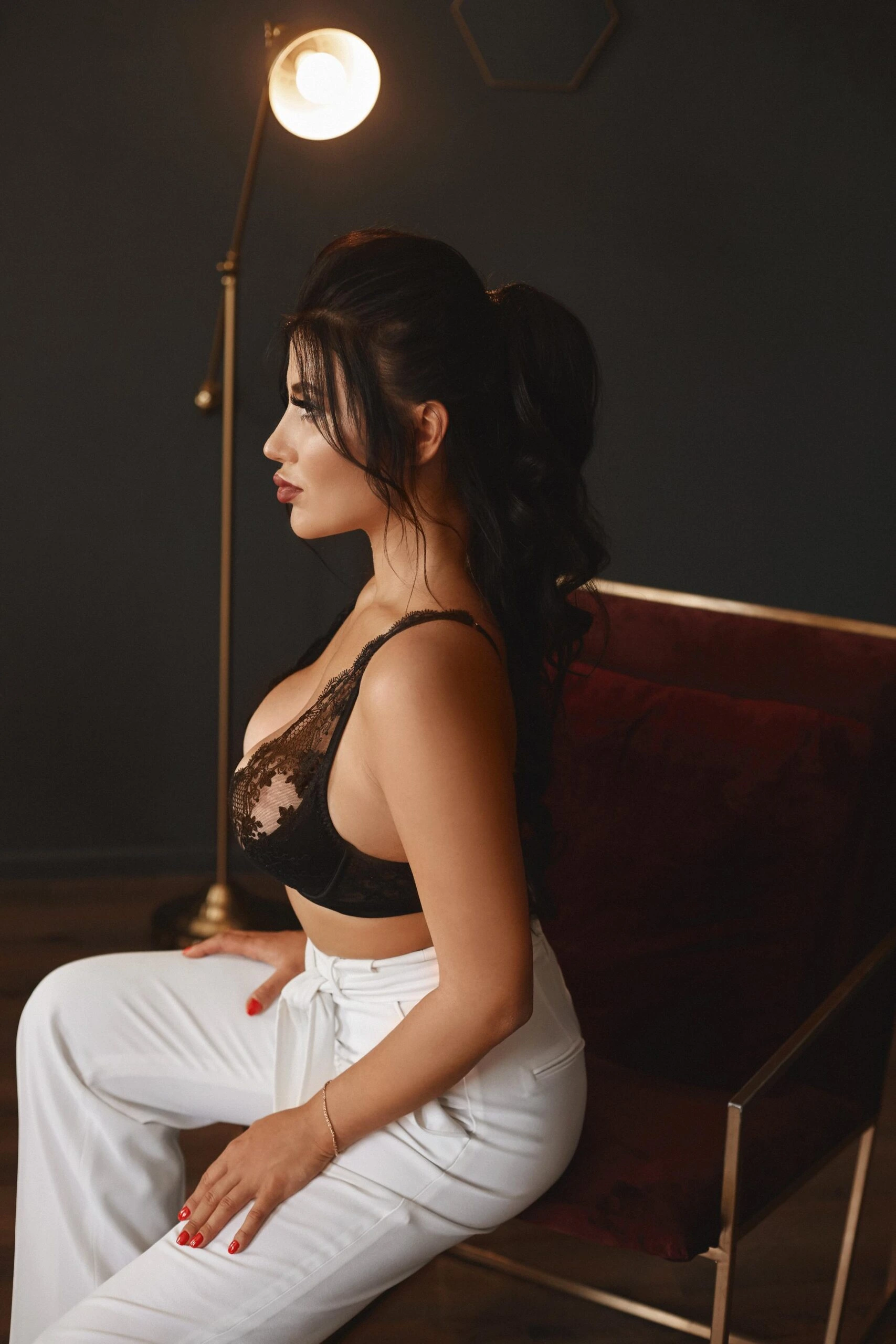 Woman in black lace top and white pants seated under a warm light in a dark room.