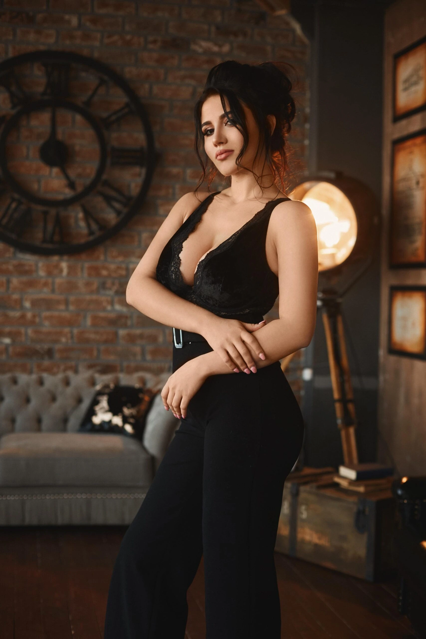 Woman in a black lace top and high-waisted pants standing against a brick wall with an industrial-style clock.