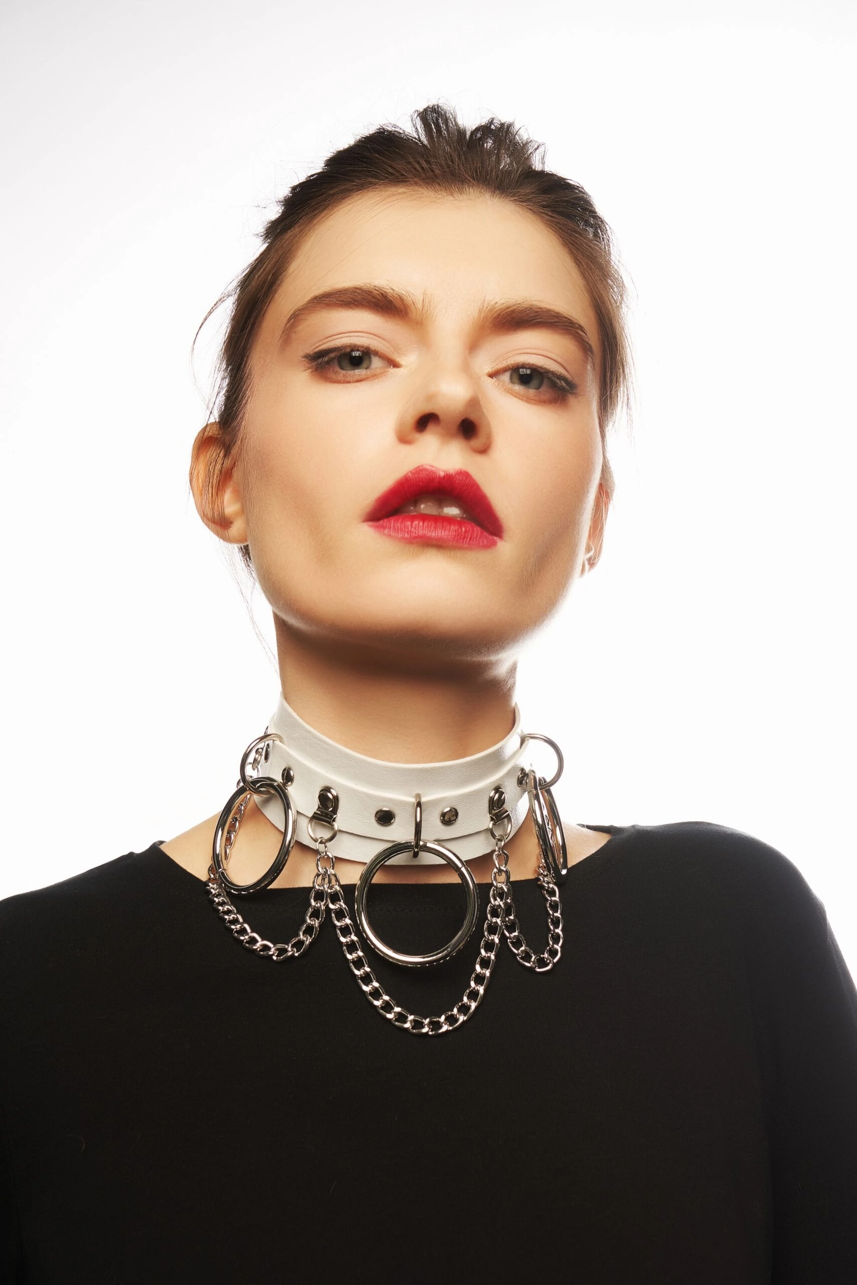 Woman with bold red lipstick wearing a white leather choker adorned with chains and rings, gazing confidently.