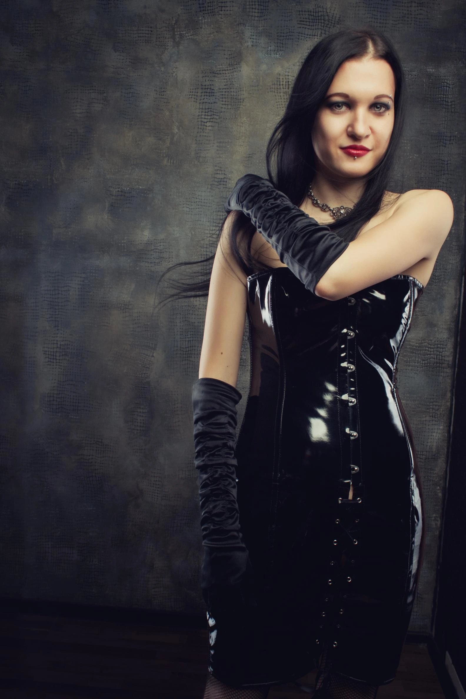Woman in a shiny black vinyl corset dress with long gloves, striking a confident pose against a textured backdrop.