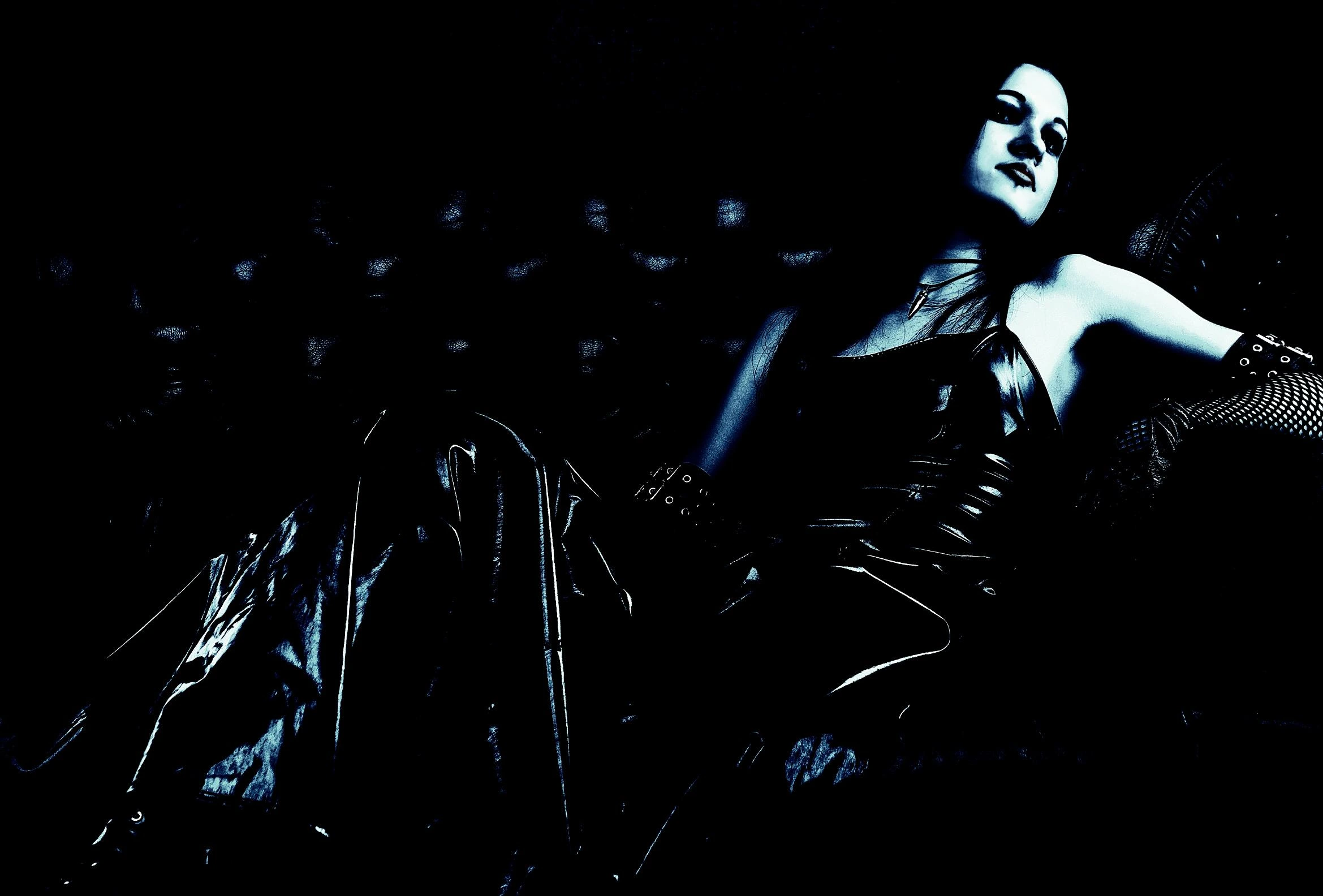 Woman reclining on a dark leather couch in a black latex dress, with dramatic lighting creating a mysterious gothic atmosphere.