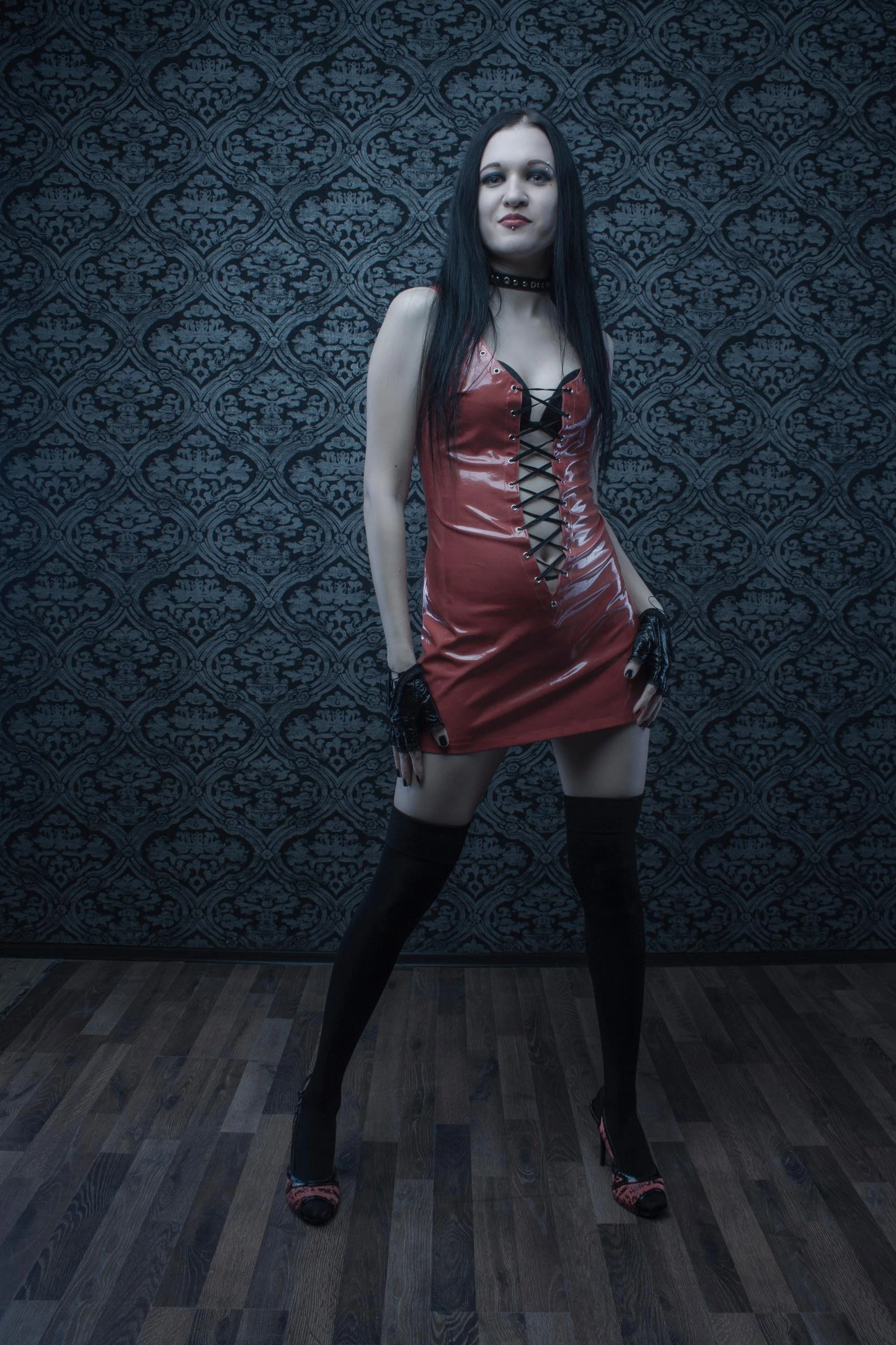 Woman in a bold red vinyl mini dress with lace-up details, styled with thigh-high stockings and black gloves.