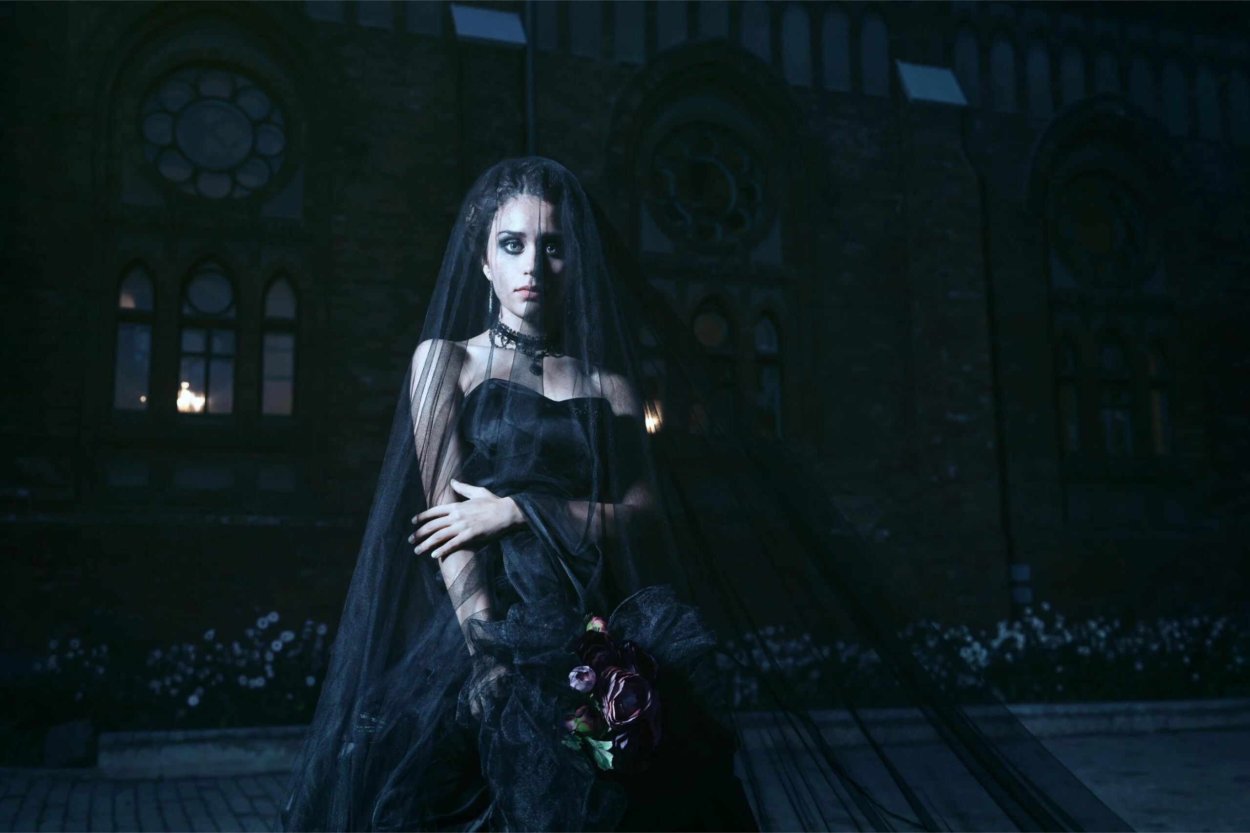 A gothic bride dressed in a black veil and flowing dress, standing solemnly in front of an old church at night, holding dark flowers.