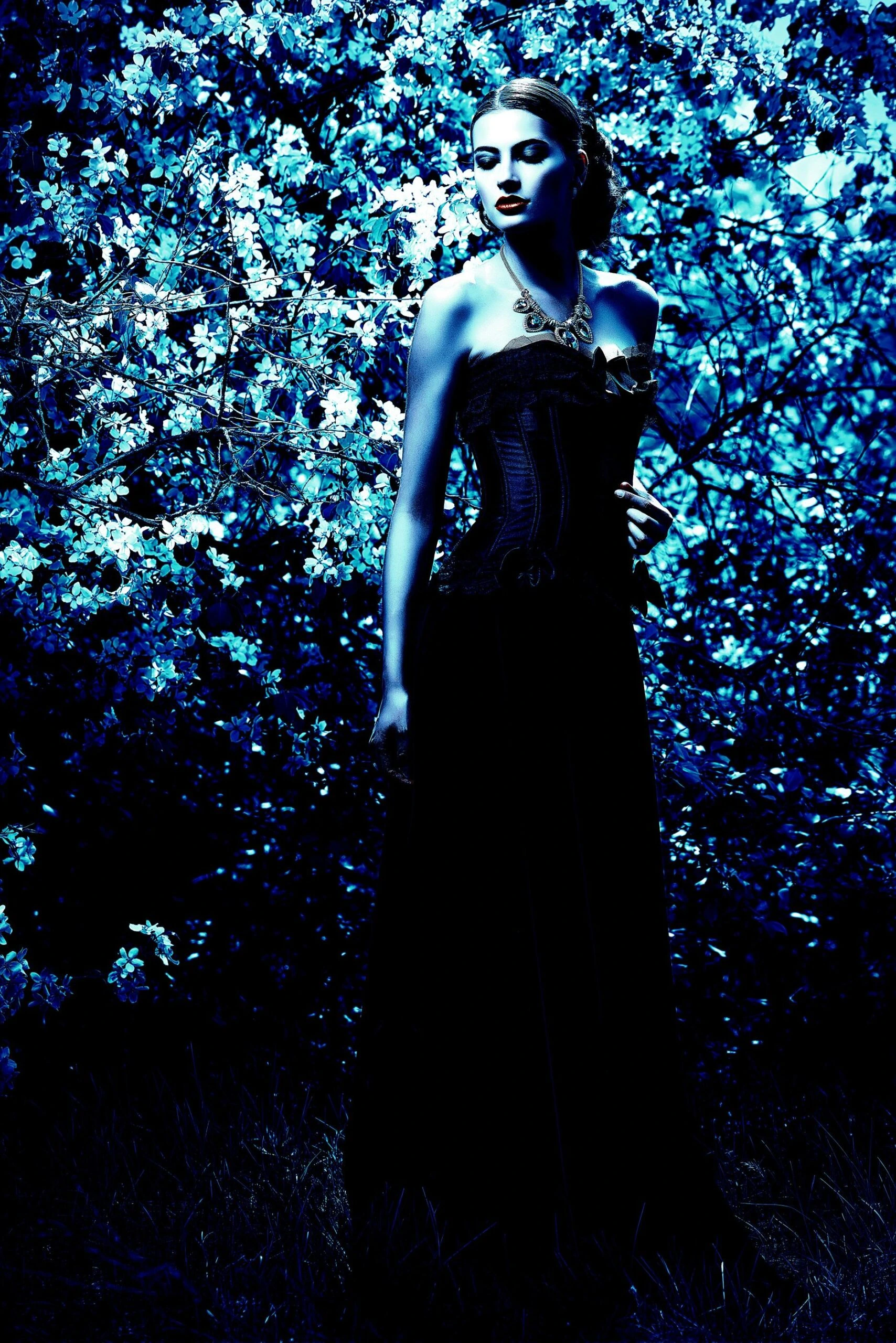 Model in a strapless black corset dress standing amidst blooming flowers under blue-tinted lighting, embodying gothic sophistication.